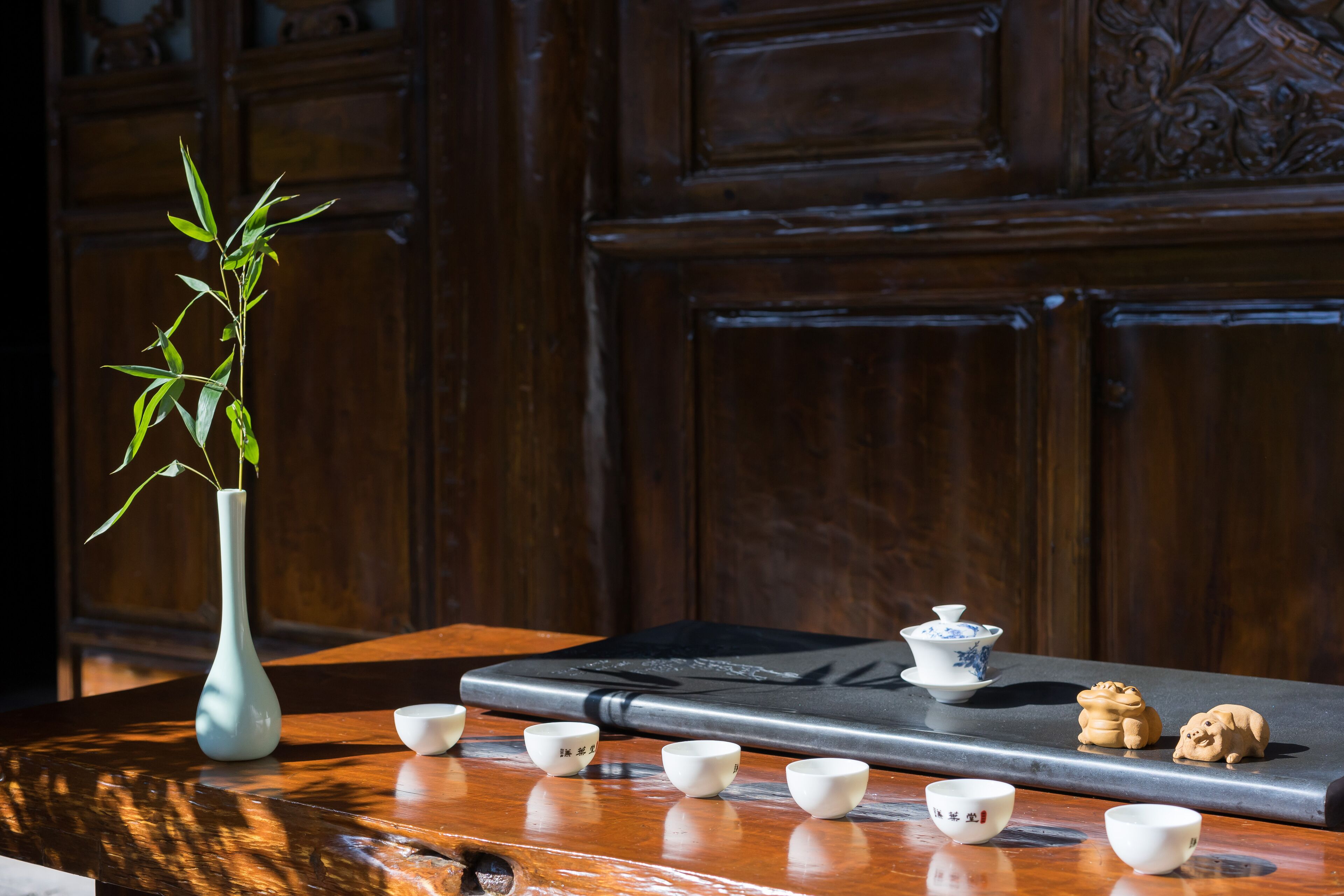Una mesa de madera con un jarrón blanco y una planta de bambú, junto con varias tazas blancas pequeñas y un hervidor de agua.