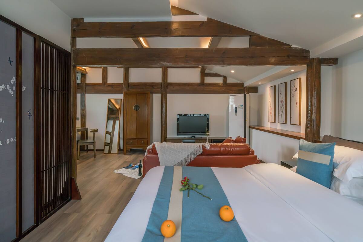 A hotel room with a wooden ceiling, a bed with a blue runner, two oranges, a rose, and a TV in the background.