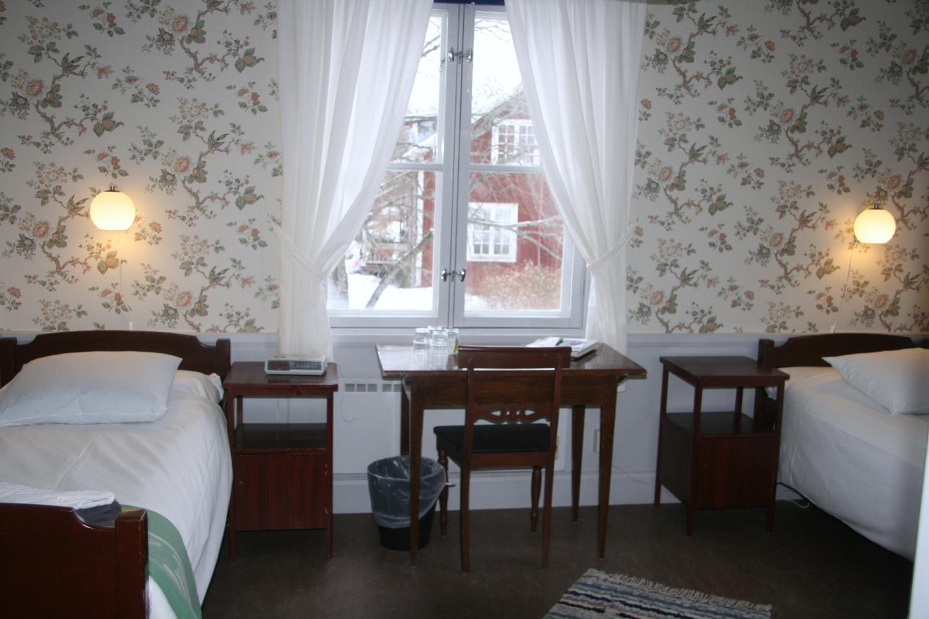 Une chambre avec deux lits, un bureau et une chaise. Il y a une fenêtre avec des rideaux, une poubelle et un tapis sur le sol.