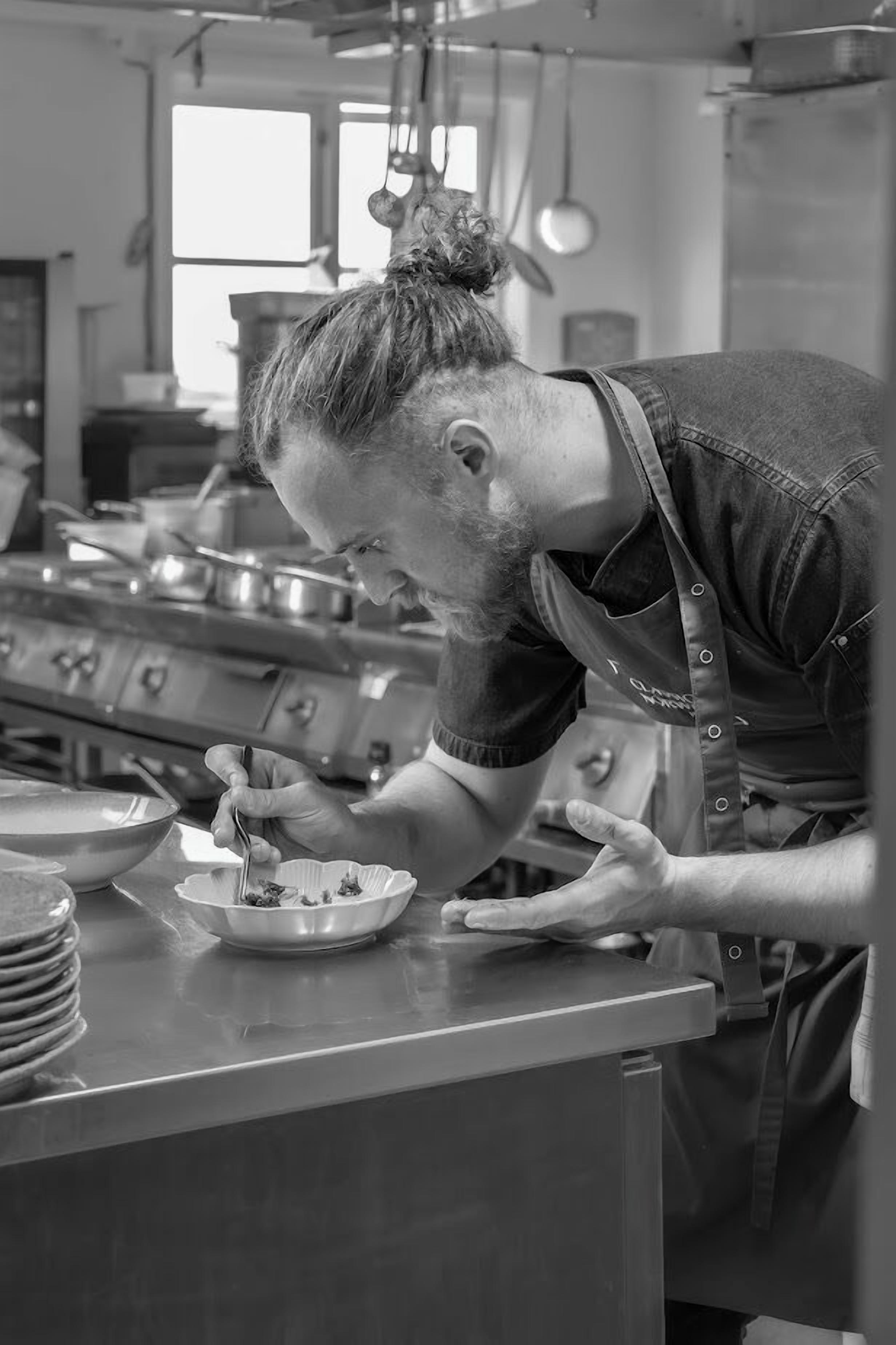 A chef in a kitchen with stainless steel appliances, a window, and hanging utensils.