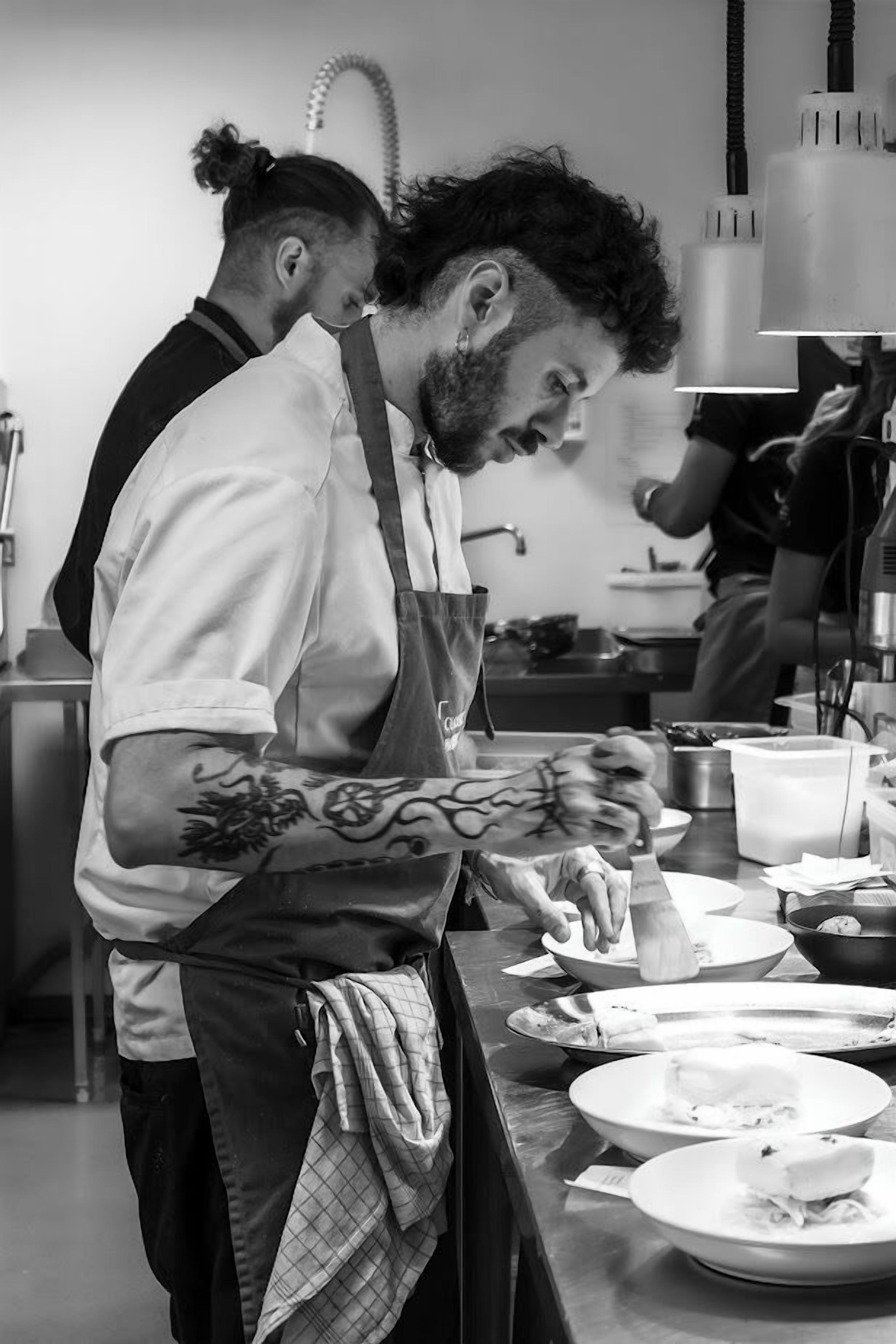 Uno chef prepara dei cibi in una cucina con piani di lavoro in acciaio inossidabile e diversi utensili da cucina.