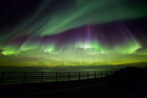 Vista nocturna de las auroras boreales sobre una carretera costera con barandilla.