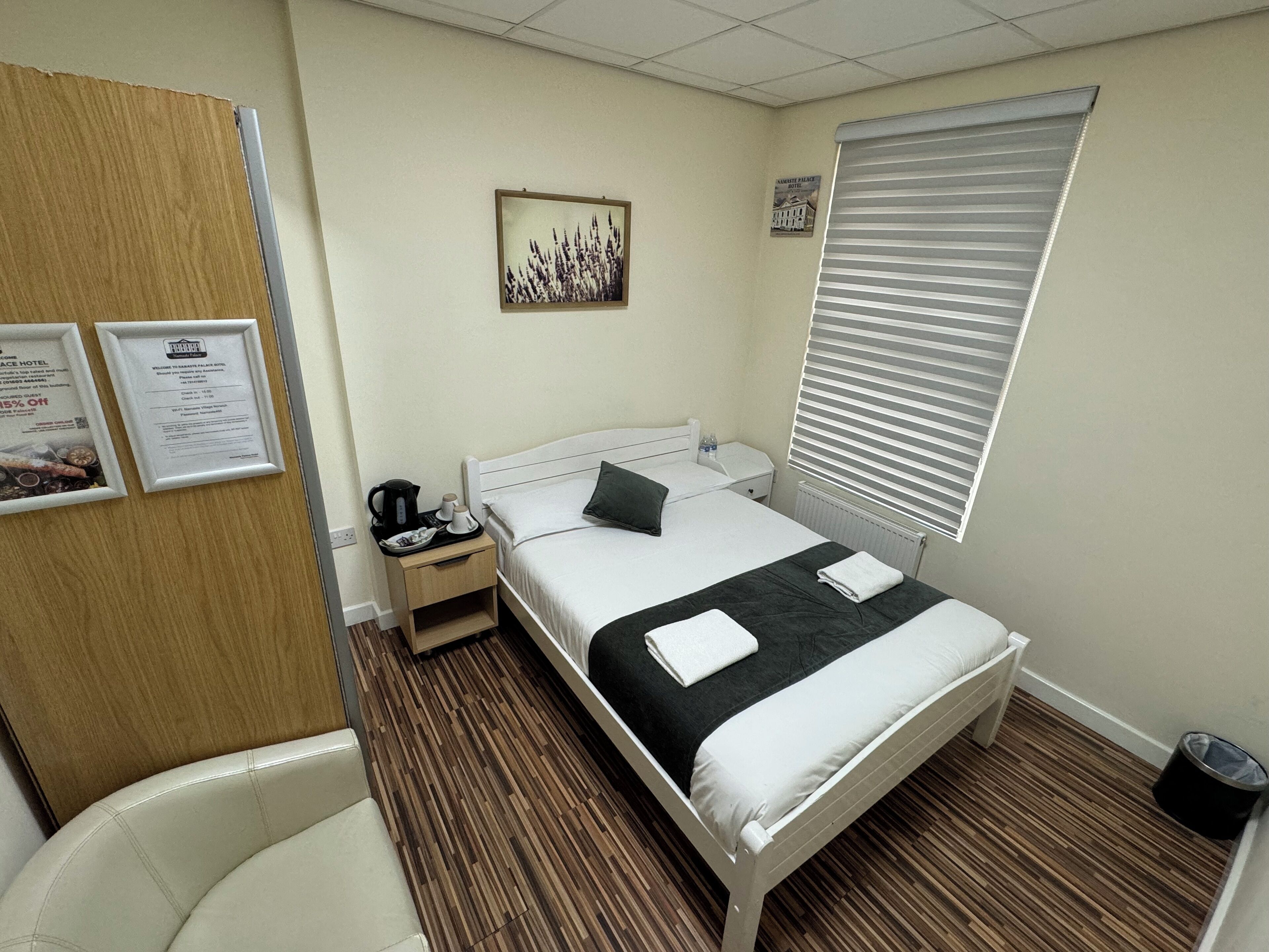 Een hotelkamer met een bed, nachtkastje, stoel, raam met zonwering en een aan de muur hangend schilderij.