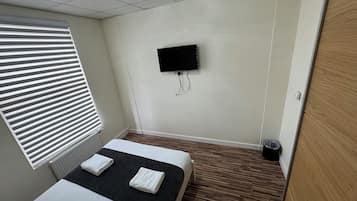 A hotel room with a bed, a TV mounted on the wall, a window with blinds, and a wooden floor.