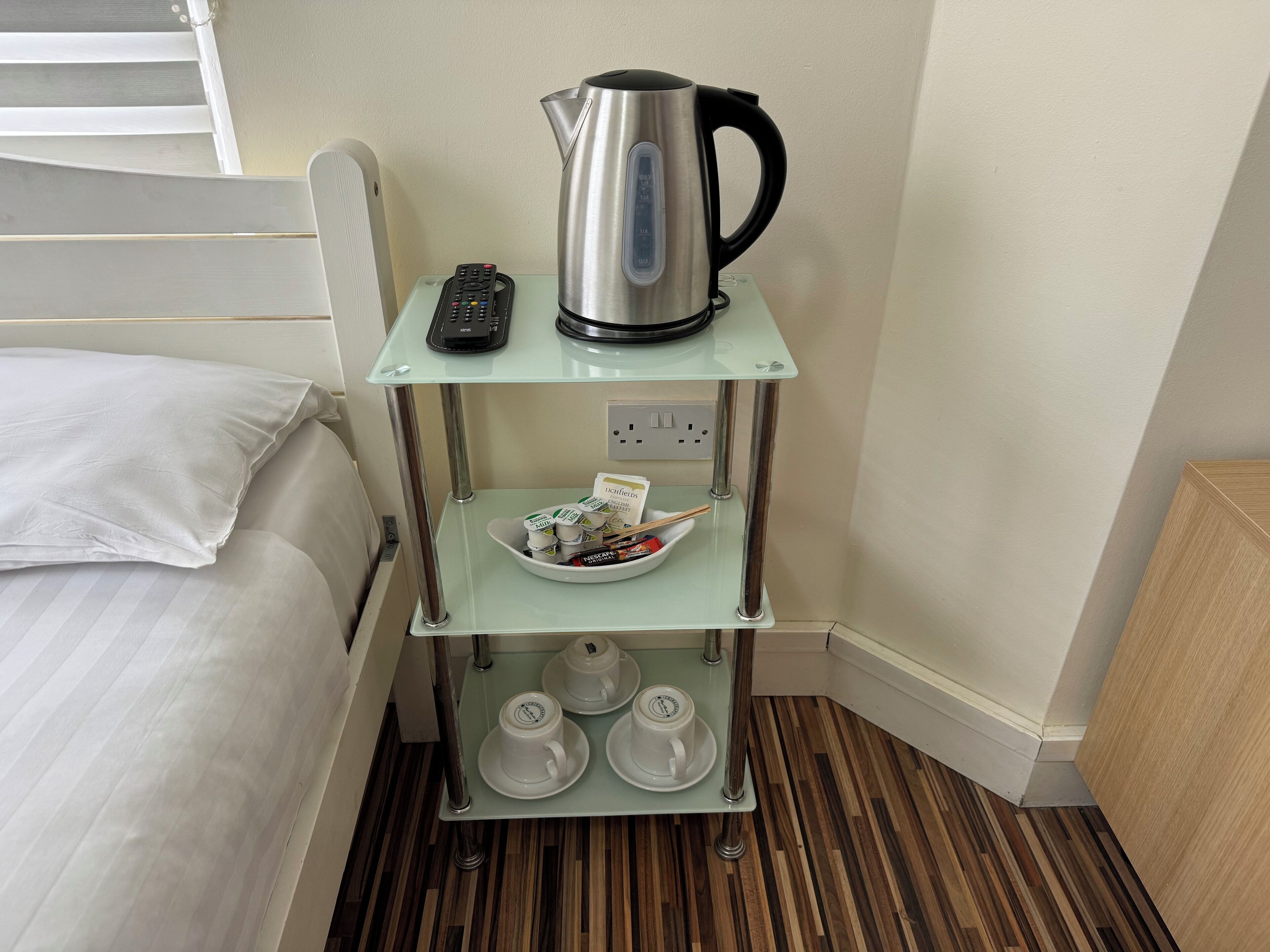 Una habitación de hotel con una cama, una pequeña mesa de vidrio con una tetera, un control remoto y un cuenco de bolsitas de té.
