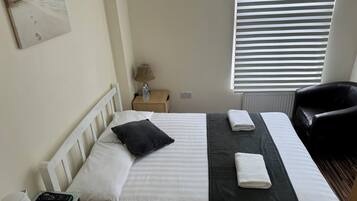 Et hotelværelse med en seng, et natbord, en lampe, en stol og et vindue med persienner.