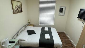 A hotel room with a bed, a bedside table with a lamp, a small shelf with a kettle and remote, a window with blinds, and a painting on the wall.