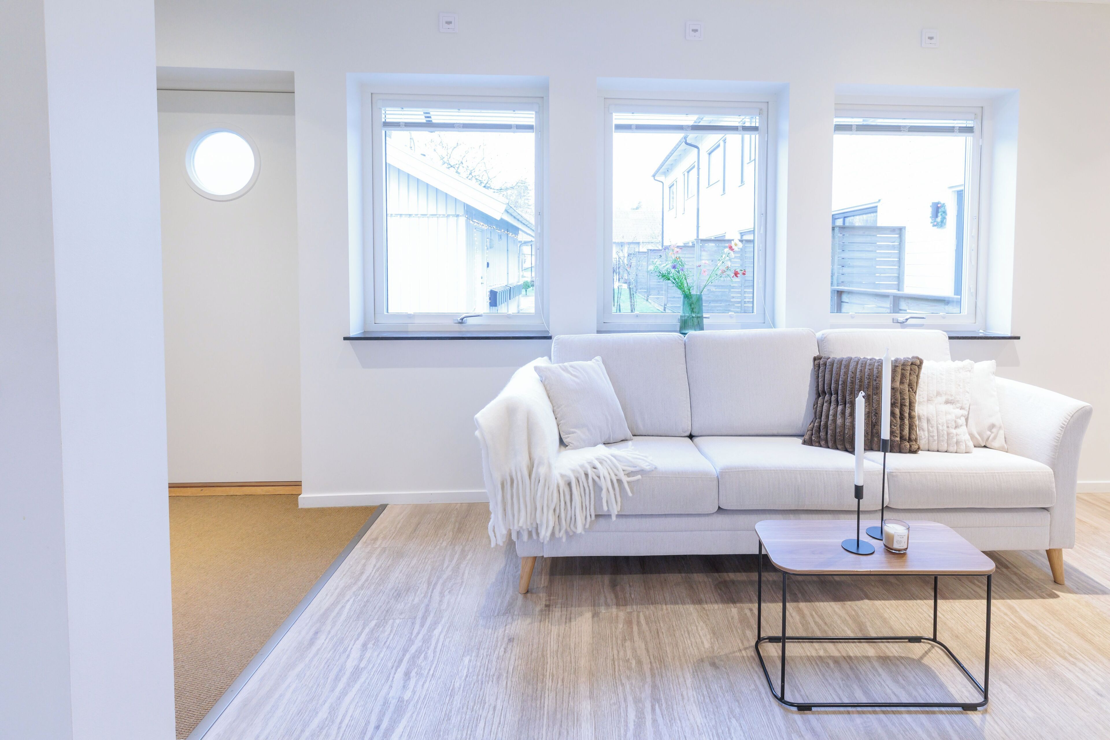 Uma sala de estar moderna com um sofá branco, uma pequena mesa lateral e três janelas com vista para o exterior.