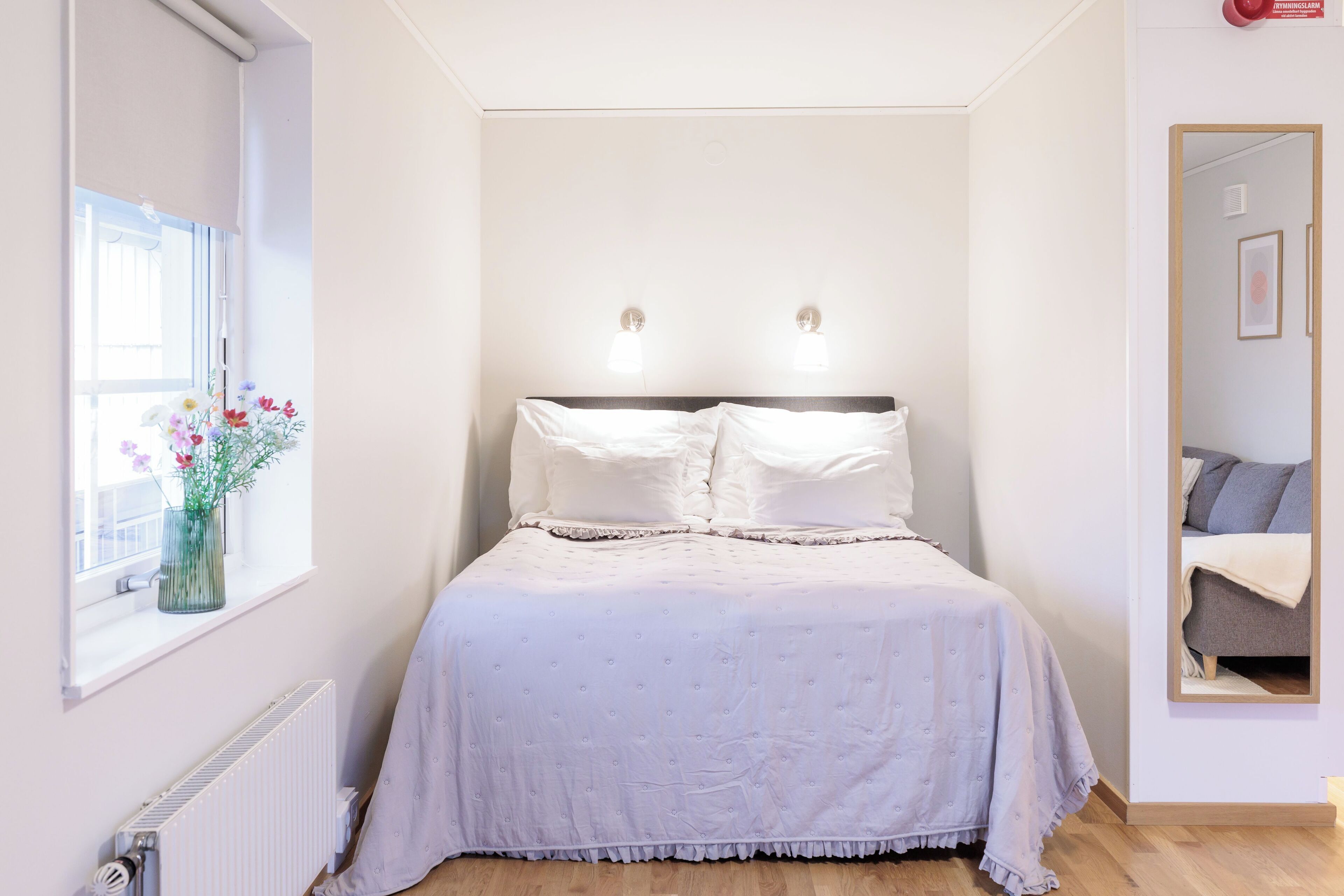 Un dormitorio con cama, una ventana llena de flores, un radiador y un espejo.