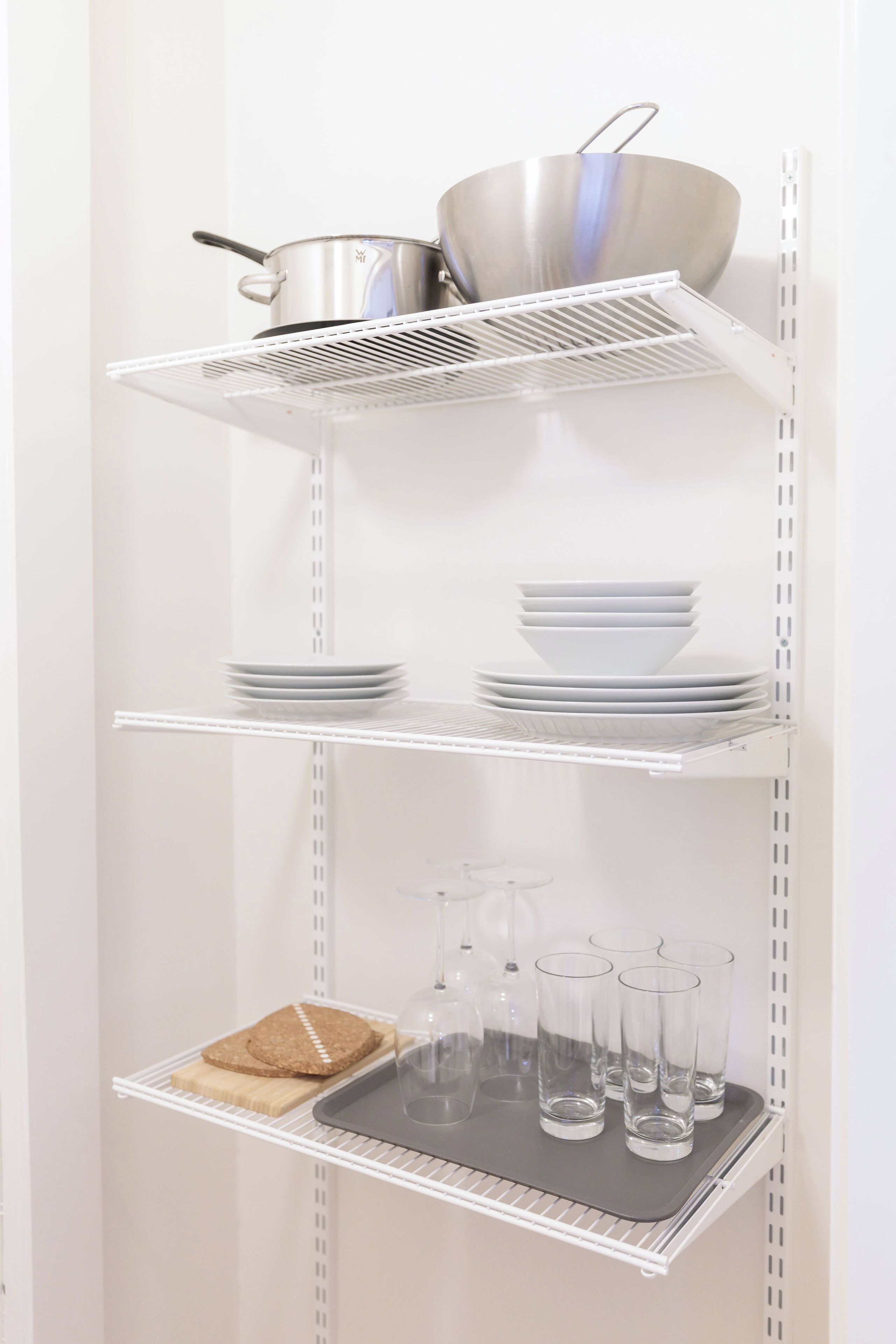 Une étagère en métal blanc contenant des ustensiles de cuisine, des assiettes, des verres et du pain.