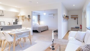 Un salon moderne et bien éclairé, avec un canapé blanc, un coin repas doté d’une table et de chaises en bois, et un espace cuisine équipé d’un four à micro-ondes et d’une machine à café.