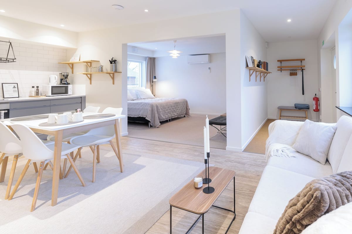 Ein modernes, helles Wohnzimmer mit einem weißen Sofa, einem Essbereich mit Holztisch und Stühlen sowie einer Küchenzeile mit Mikrowelle und Kaffeemaschine.