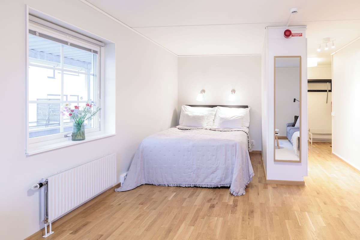 Ein Schlafzimmer mit einem Bett, einem Fenster mit Blumen, einem Heizkörper und einem Spiegel.