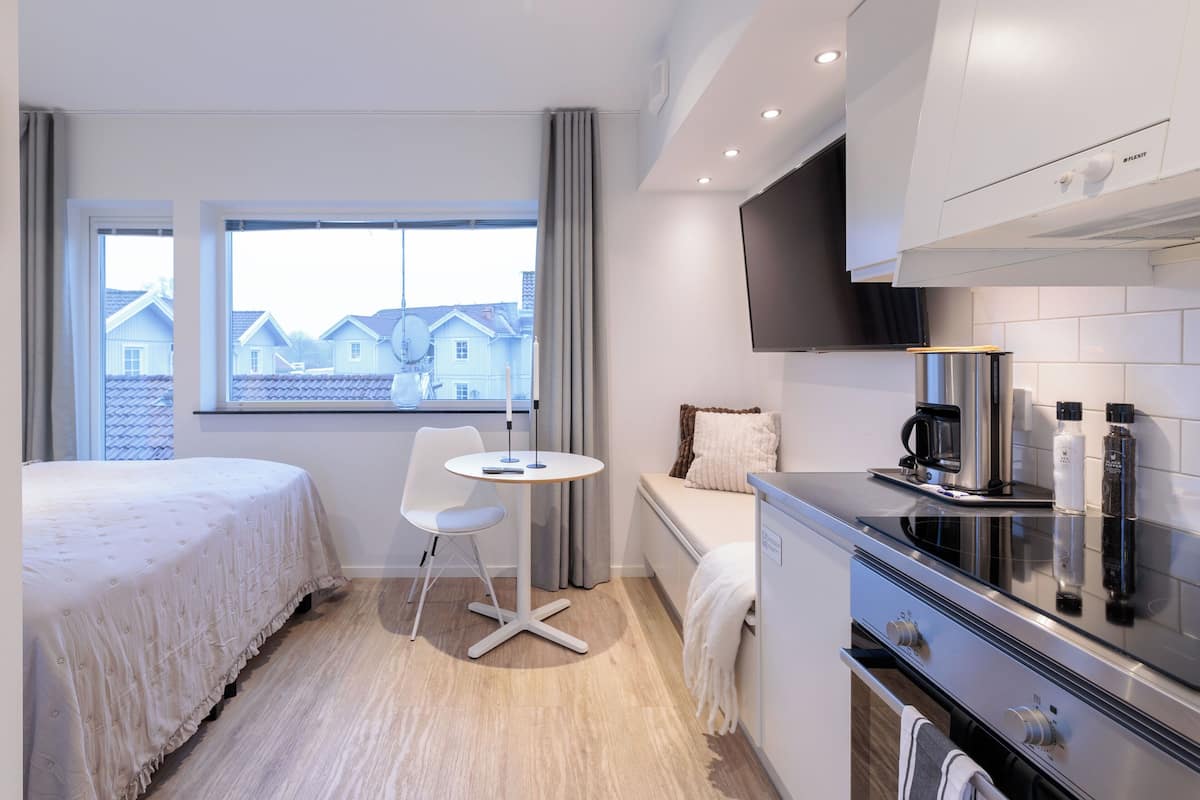 Ein modernes Hotelzimmer mit Bett, kleinem Tisch, Stuhl, Sofa und Küchenzeile inklusive Herd. Ein Fenster bietet Blick auf Häuser.