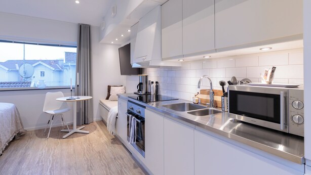 Une cuisine moderne équipée d’appareils électroménagers en acier inoxydable, un coin repas avec une table et des chaises blanches, et une fenêtre donnant sur des maisons.