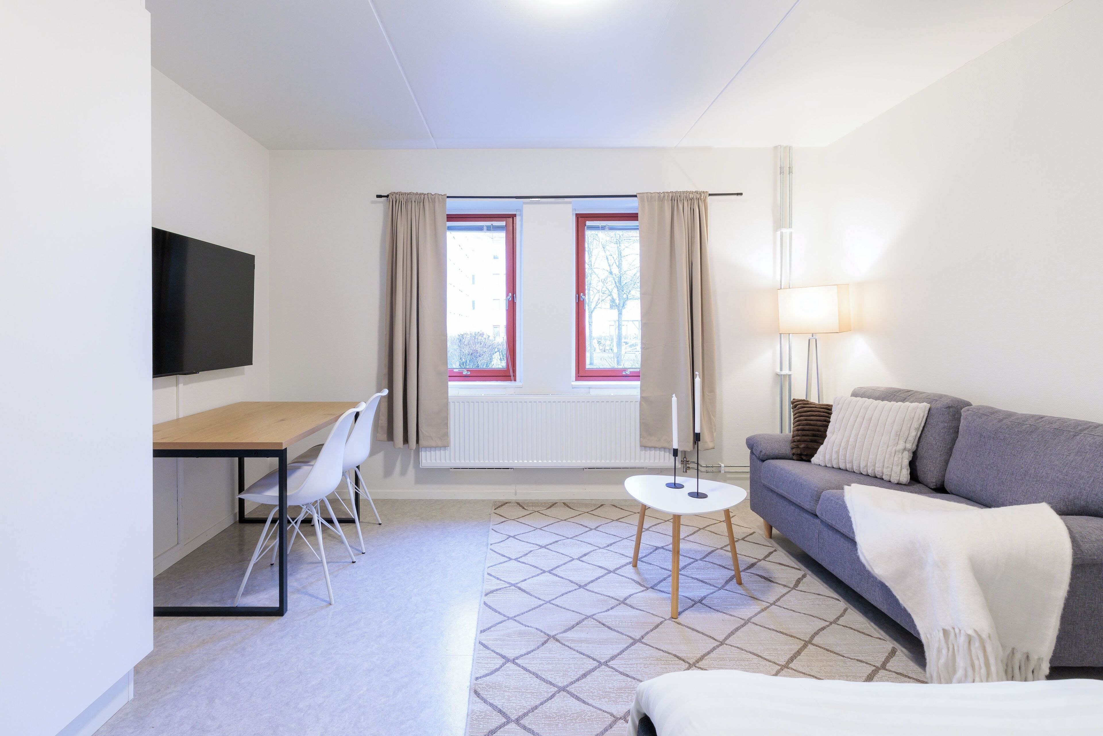 A modern room with a sofa, a dining table with chairs, a TV mounted on the wall, and a window with curtains.