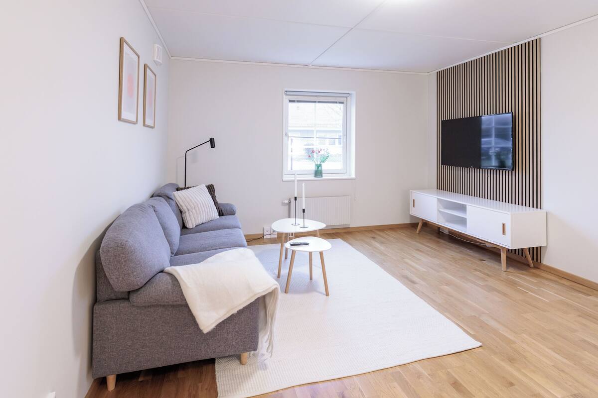 Ein modernes Wohnzimmer mit grauem Sofa, einem weißen Teppich, einem kleinen weißen Tisch, einer weißen TV-Konsole und einem Fenster mit Jalousien.