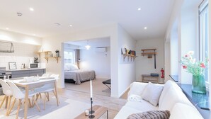 Un salon moderne avec un canapé blanc, un coin repas avec une table et des chaises blanches, et une cuisine en arrière-plan.