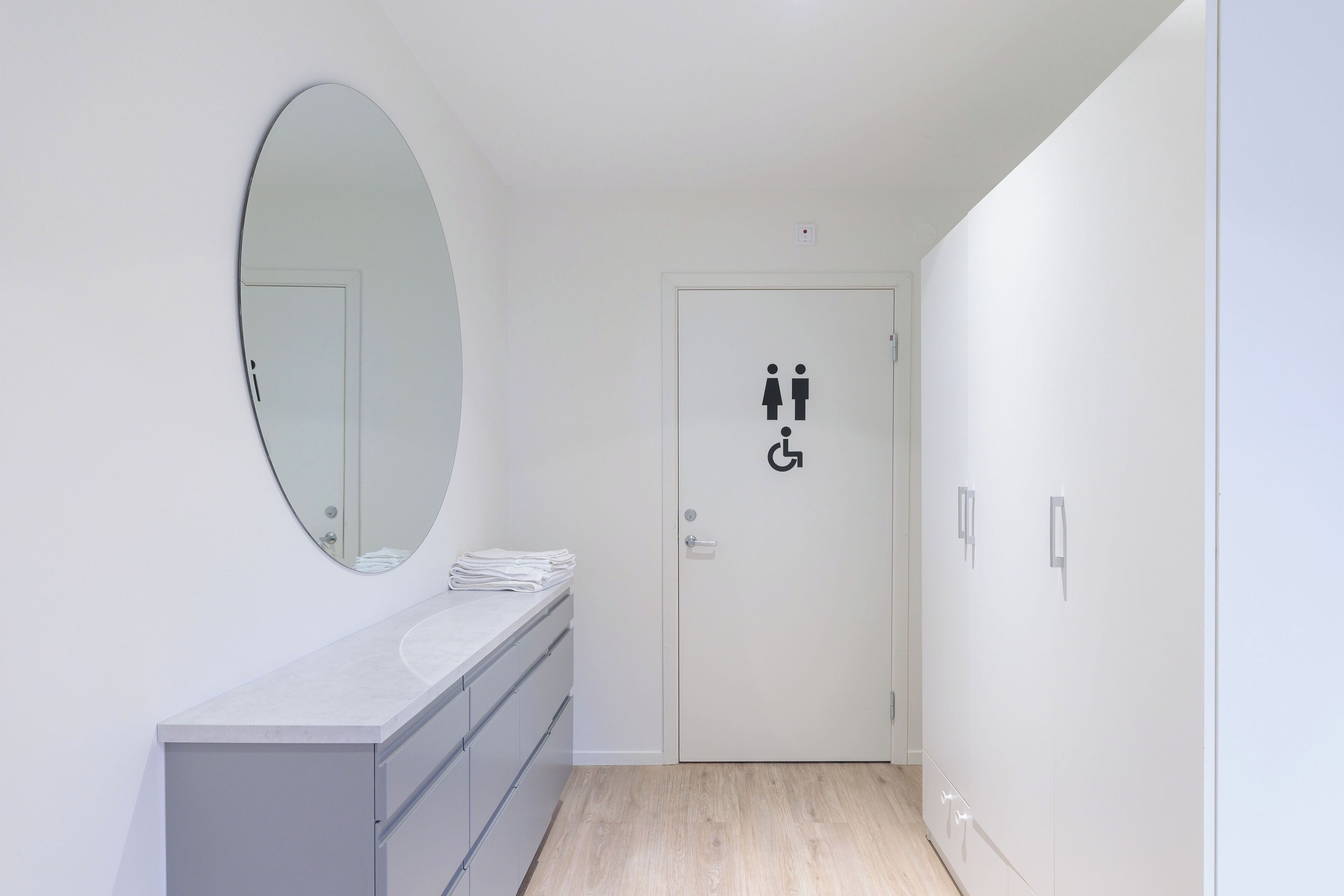 A bathroom with a white vanity, a round mirror, and a door with a gender-neutral symbol.