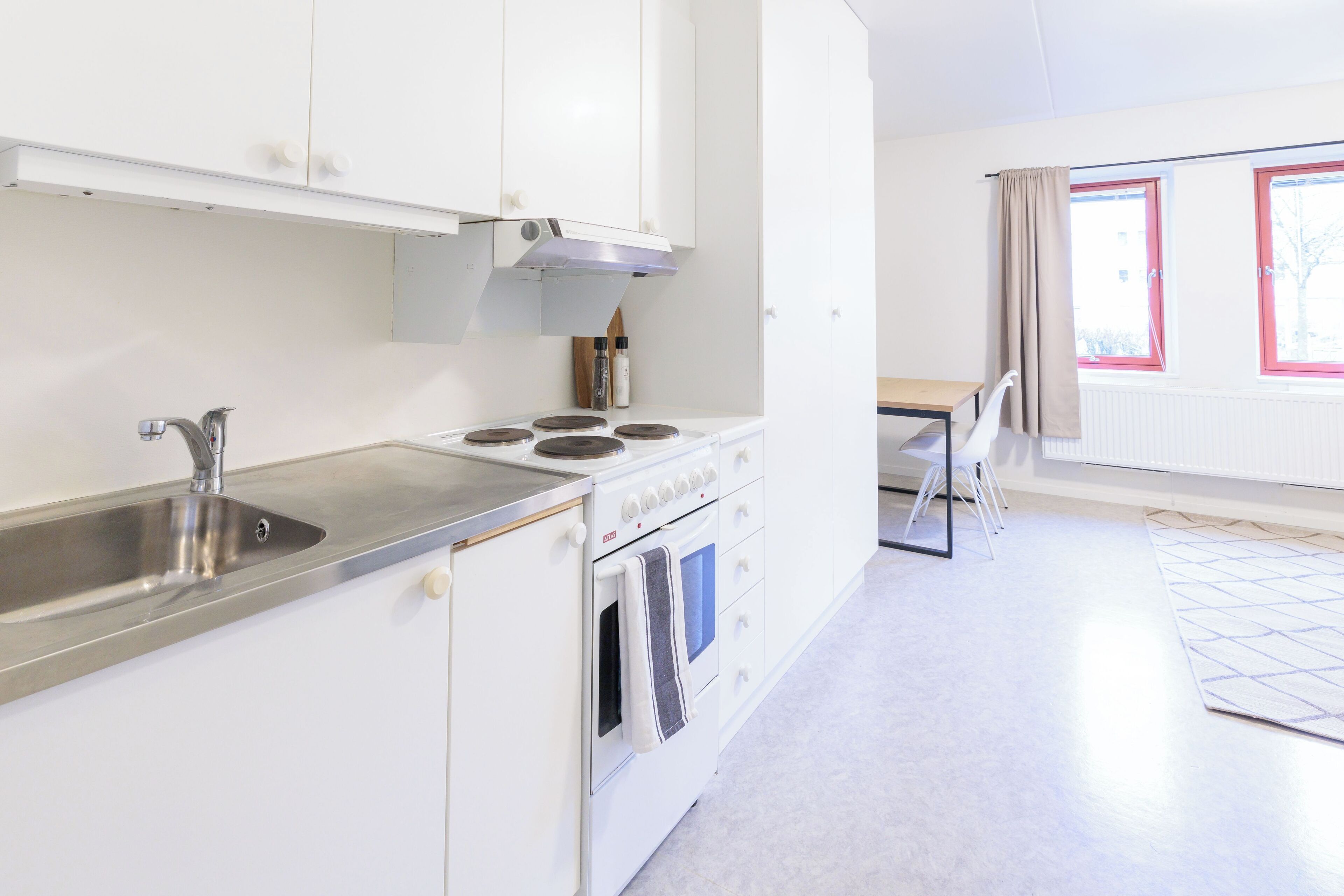 Una cocina moderna con armarios blancos, un lavamanos de acero inoxidable, una estufa y un horno. Hay una zona de comedor con una mesa y sillas.