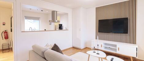 Un salon moderne comprenant un canapé, deux petites tables, un téléviseur à écran plat et un coin cuisine avec une cuisinière et une fenêtre.