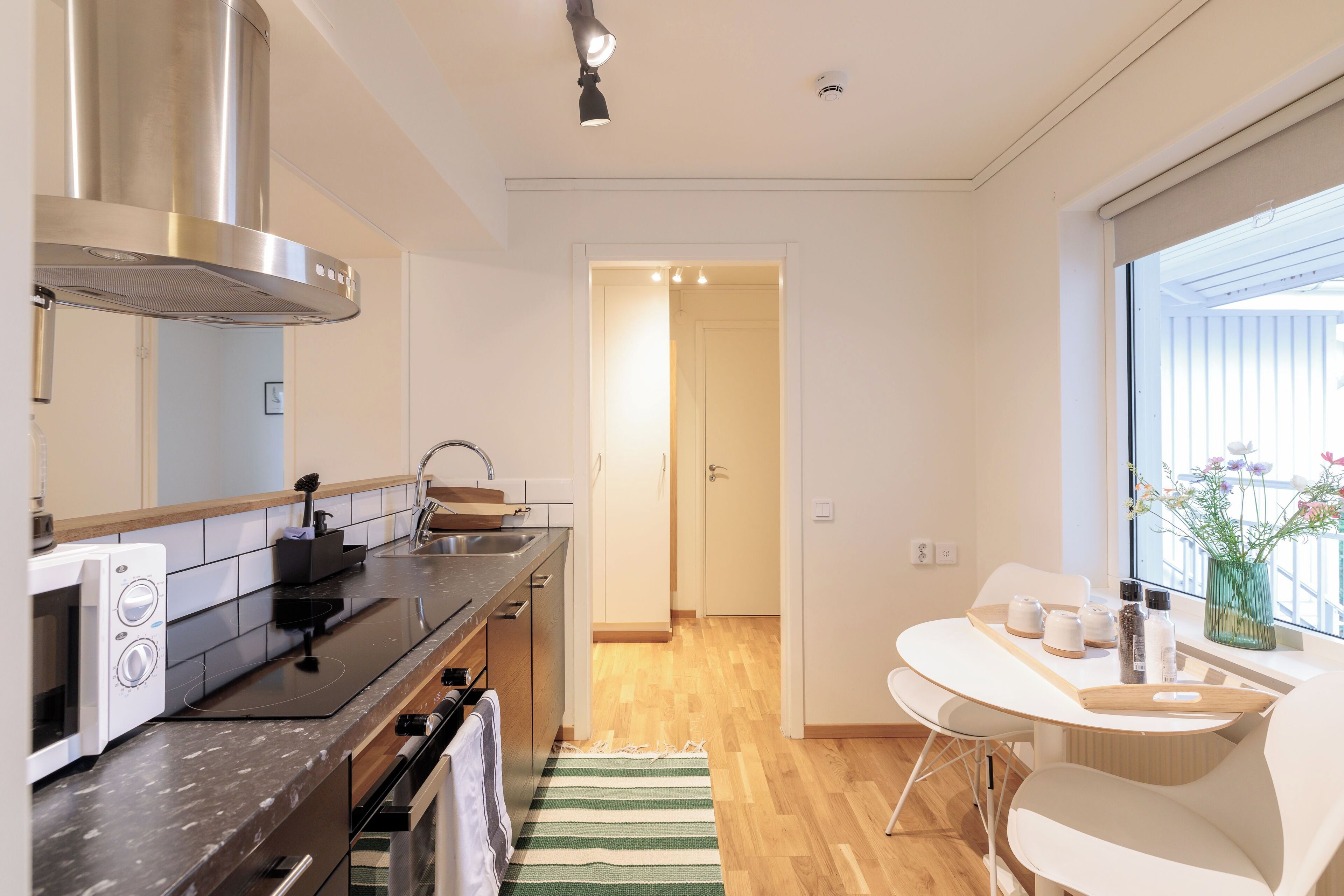 A modern kitchen with a stove, microwave, and stainless steel range hood. There is a dining area with a table and chairs, and a window with a view of the outdoors.
