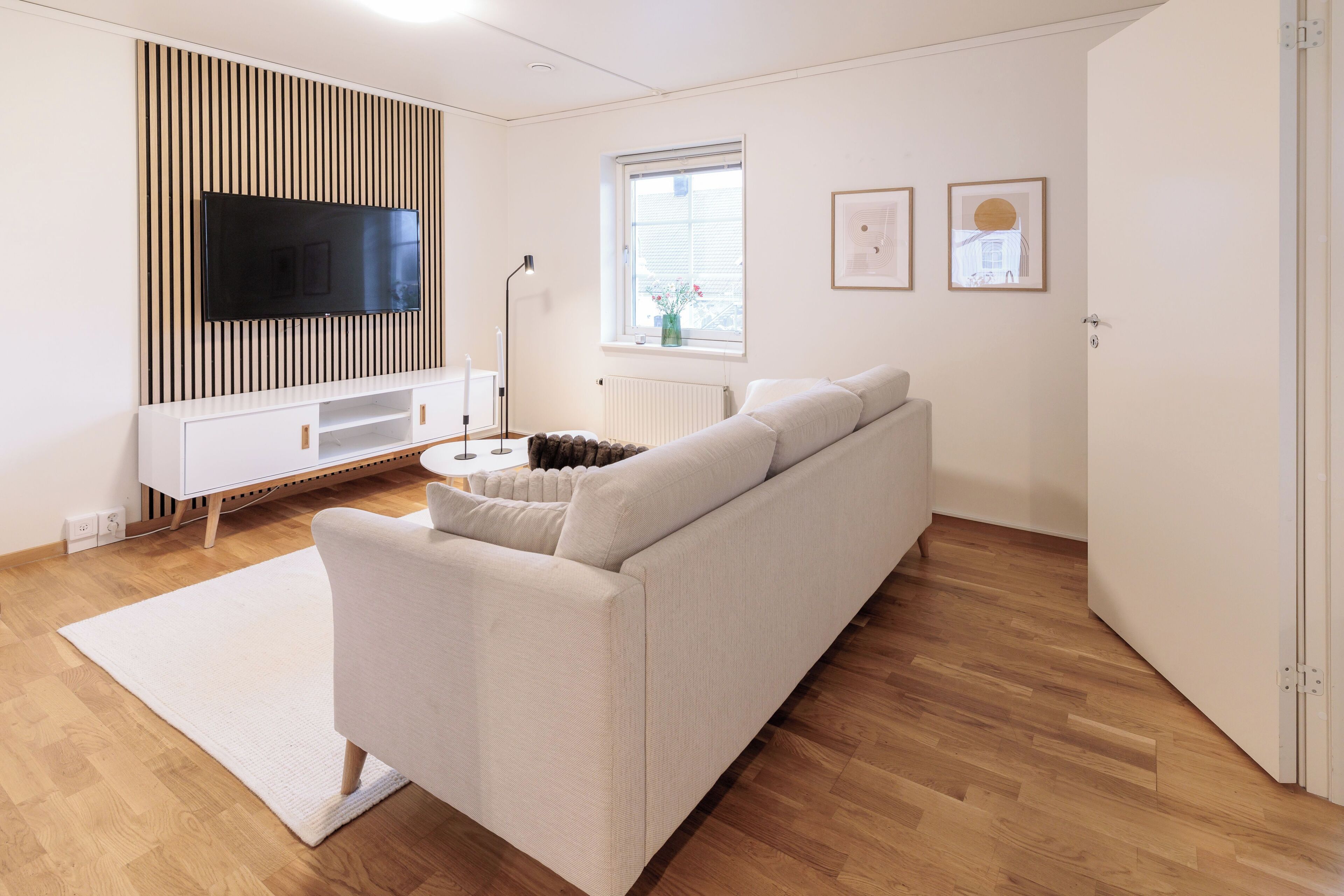 A modern living room with a white sofa, a flat-screen TV mounted on a wooden panel, a white radiator, a window with blinds, and framed pictures on the wall.