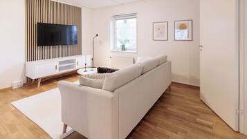 A modern living room with a white sofa, a flat-screen TV mounted on a wooden panel, a white radiator, a window with blinds, and framed pictures on the wall.