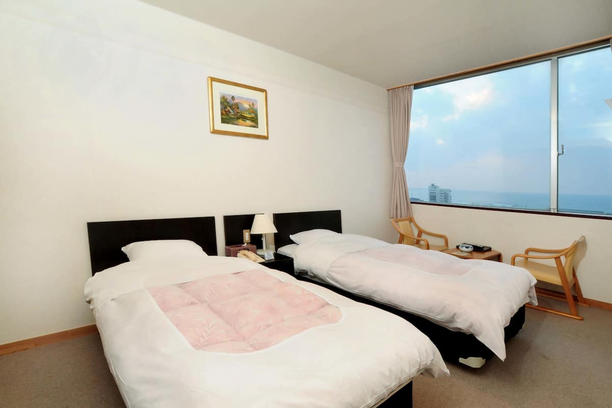 Ett hotellrum med två sängar, ett litet bord, en stol, ett fönster med utsikt och en inramad tavla på väggen.