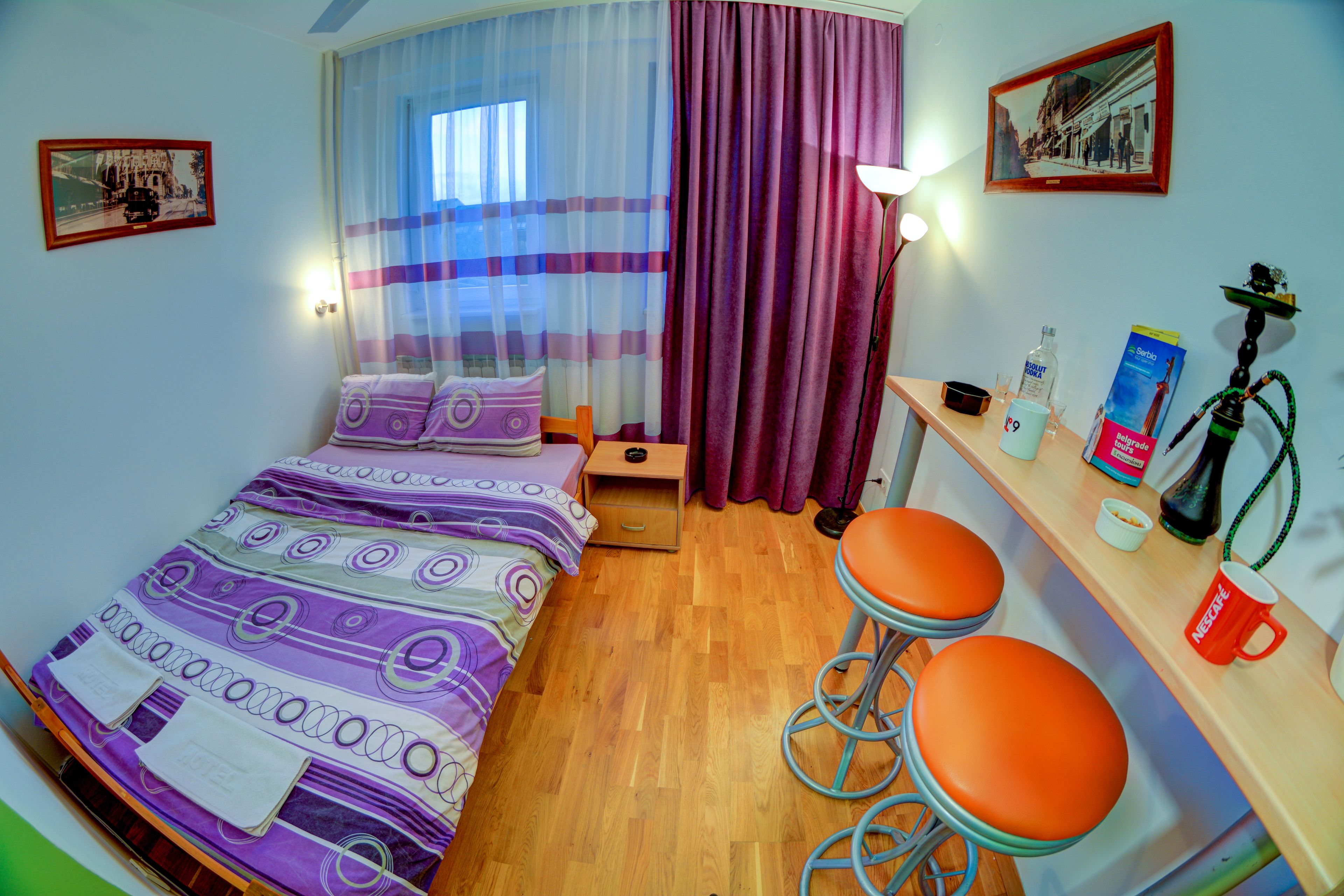 Une chambre d’hôtel de taille modeste, comprenant un lit, une petite table avec une chaise, un narguilé et une fenêtre avec des rideaux.