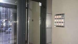 Un ascensor con puerta de cristal, panel metálico con una rejilla de luces y un pequeño cartel en la pared.