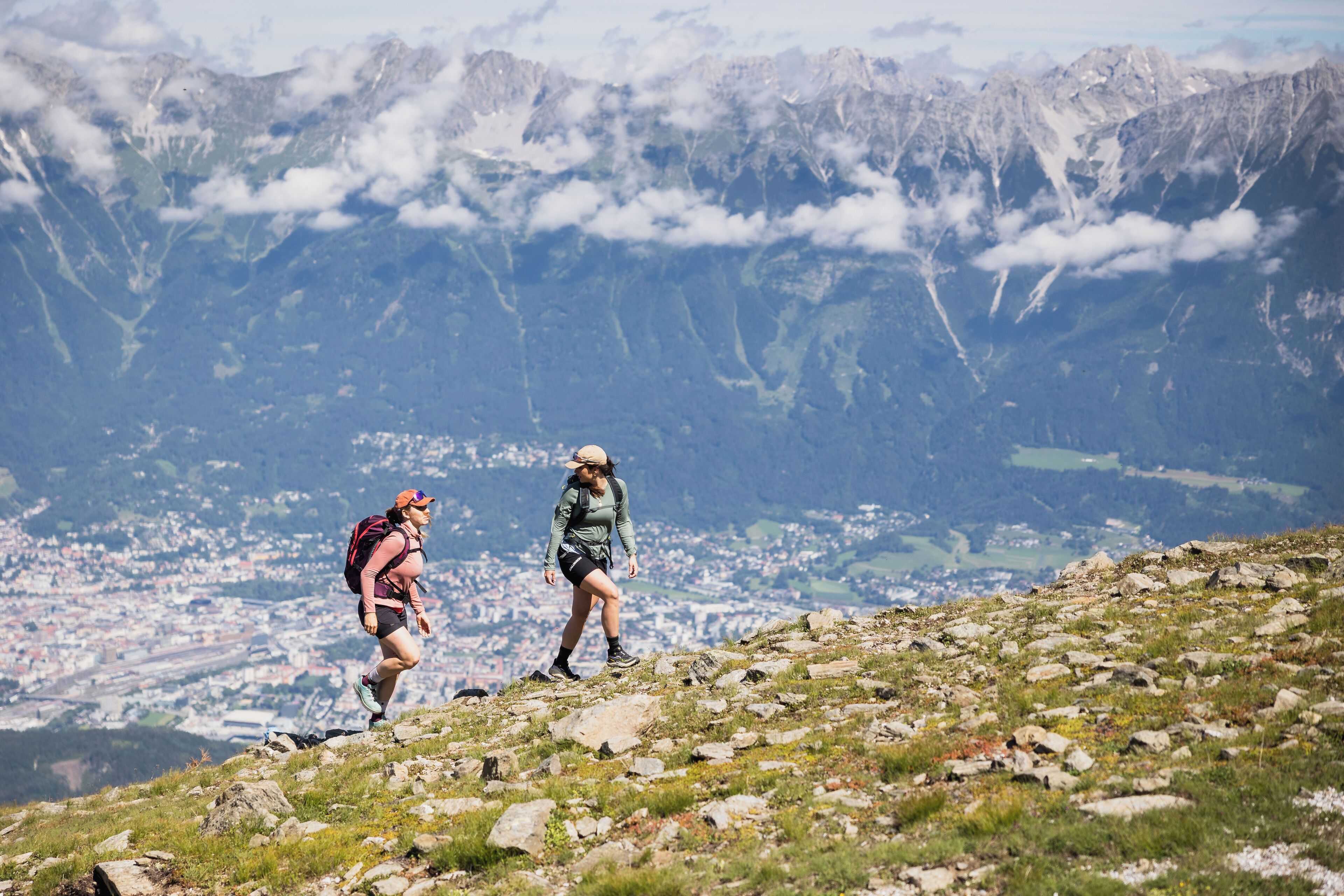 A hiker on a rocky mountain path with a city and mountains in the background.