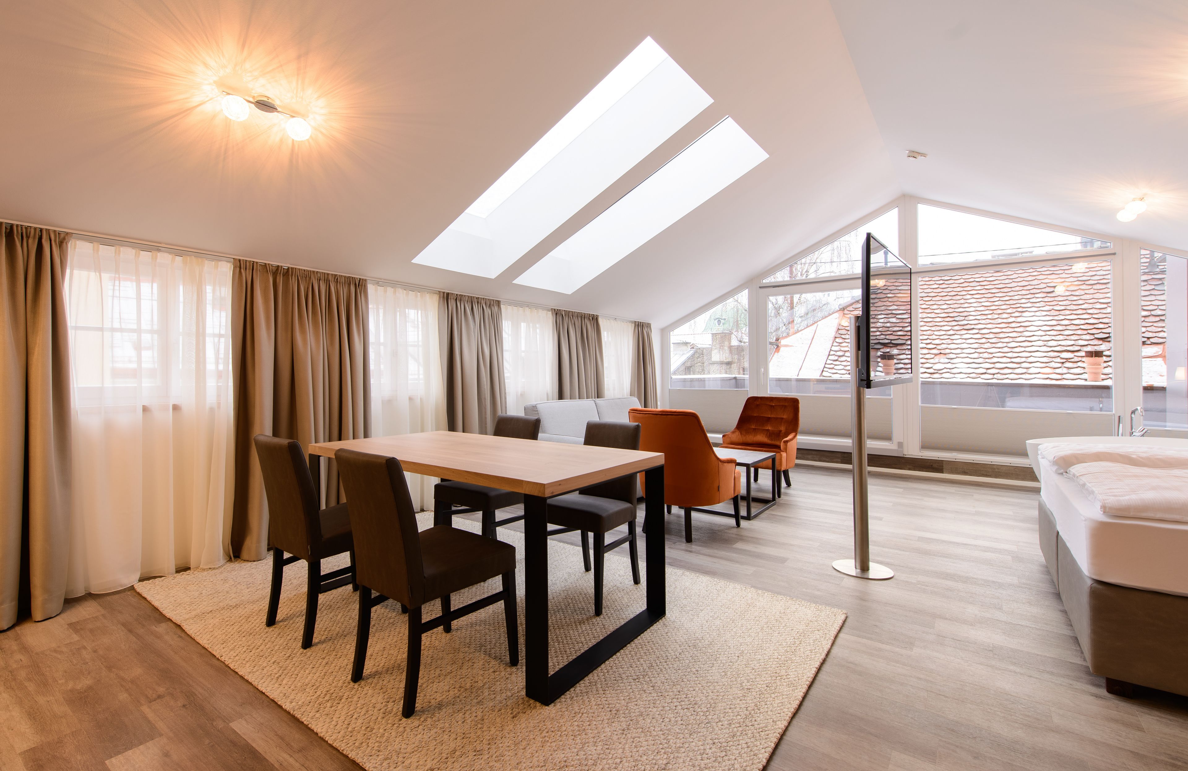 A modern room with a wooden dining table, chairs, a sofa, and a bed. There are large windows and skylights providing natural light.