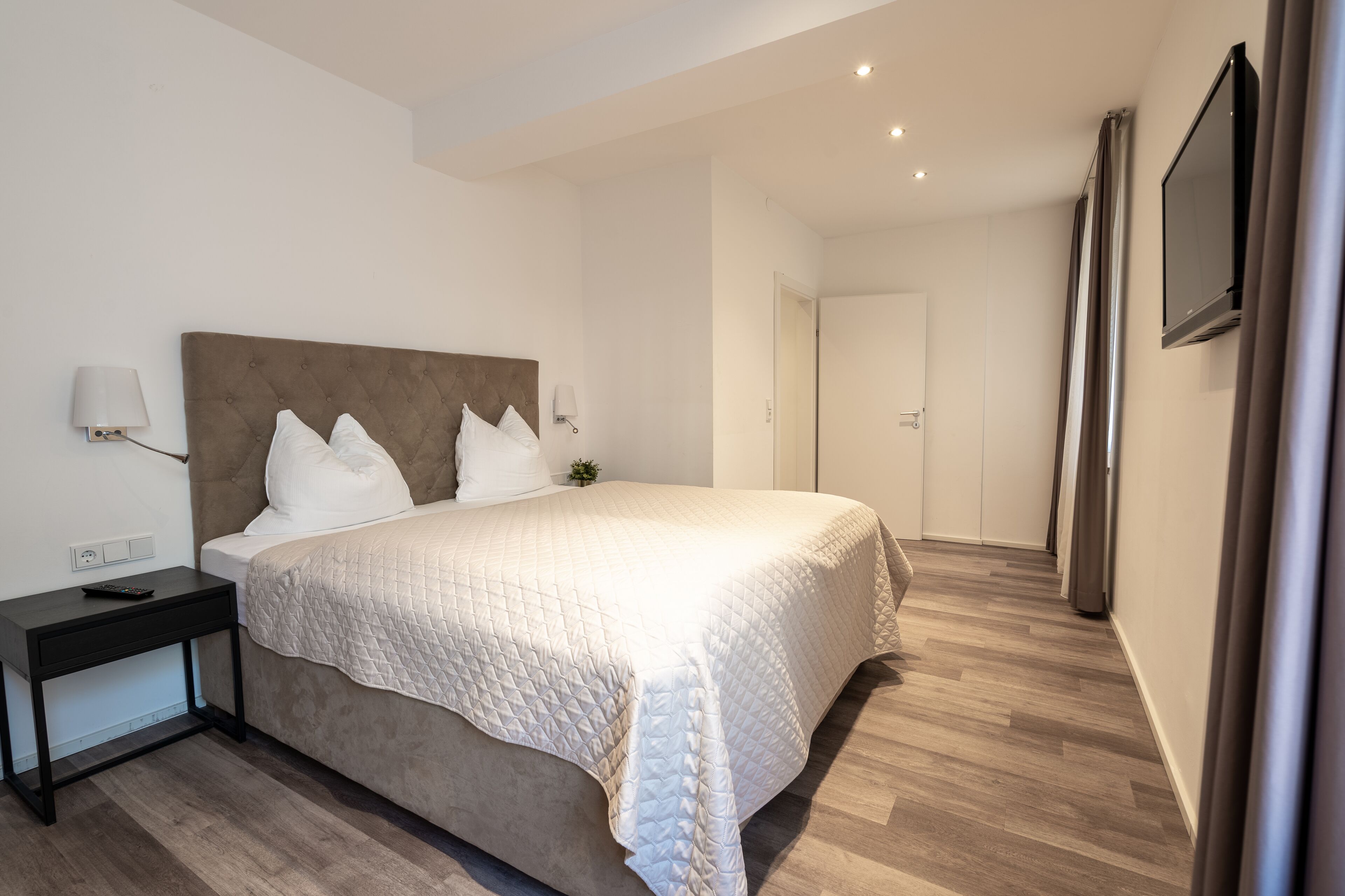 Une chambre à coucher moderne, dotée d’un grand lit, d’une table de chevet, d’un téléviseur fixé au mur et d’une porte donnant sur une autre pièce.