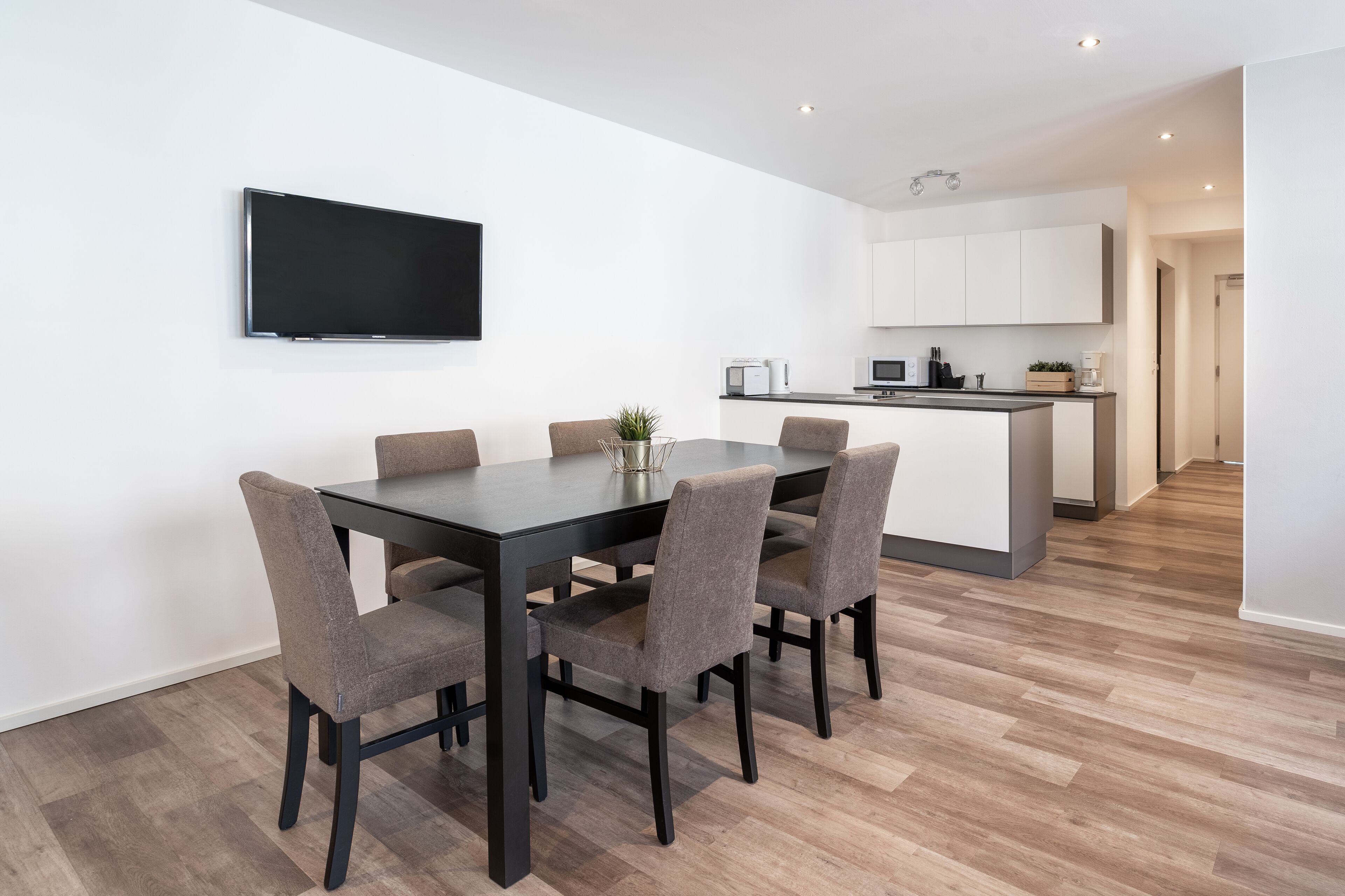 Een moderne keuken met witte kasten, een flatscreen televisie boven een eettafel met vier stoelen, en een klein kookeiland met apparatuur.
