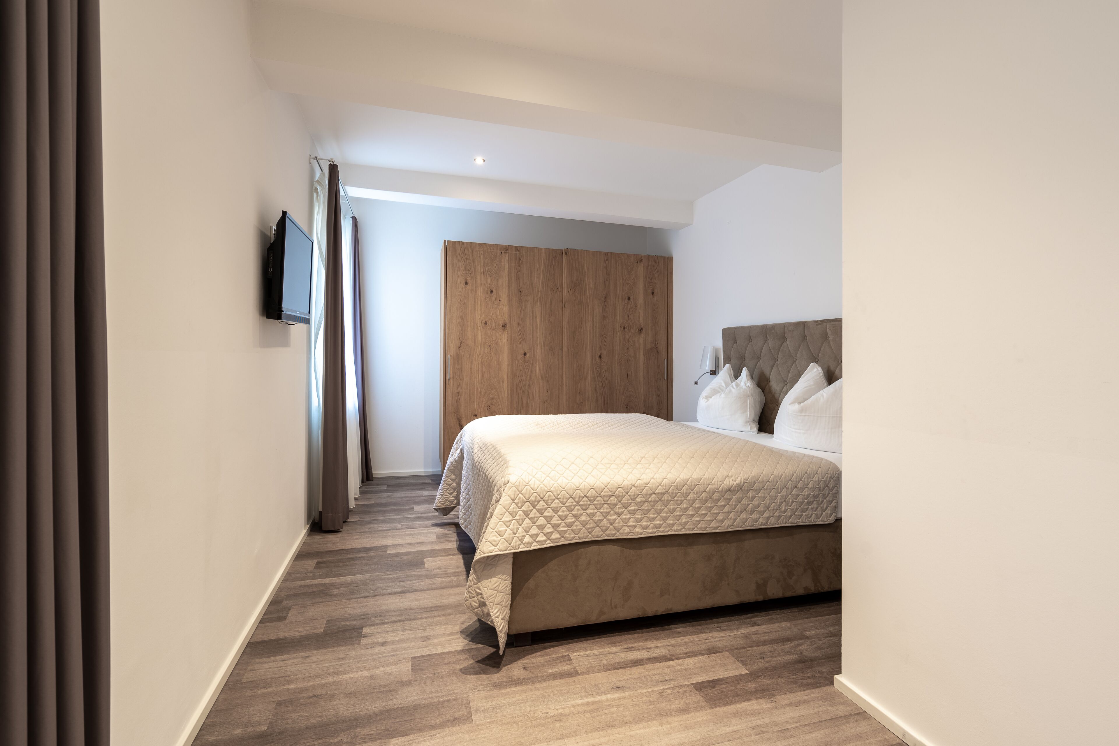 A modern bedroom with a large bed, a wooden wardrobe, and a flat-screen TV mounted on the wall.