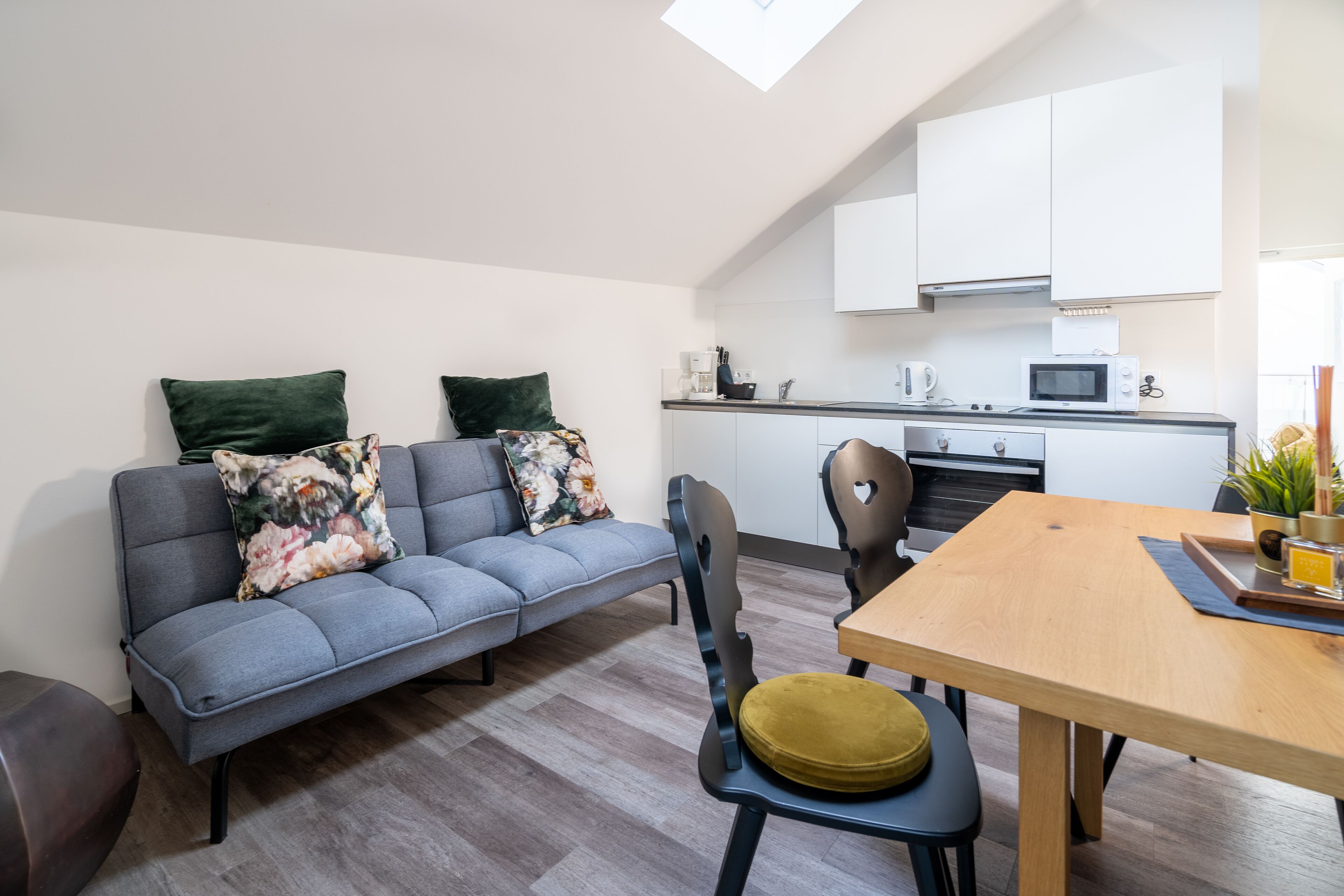 A modern living space with a grey sofa, a wooden dining table, and a kitchen area with white cabinets and a microwave.