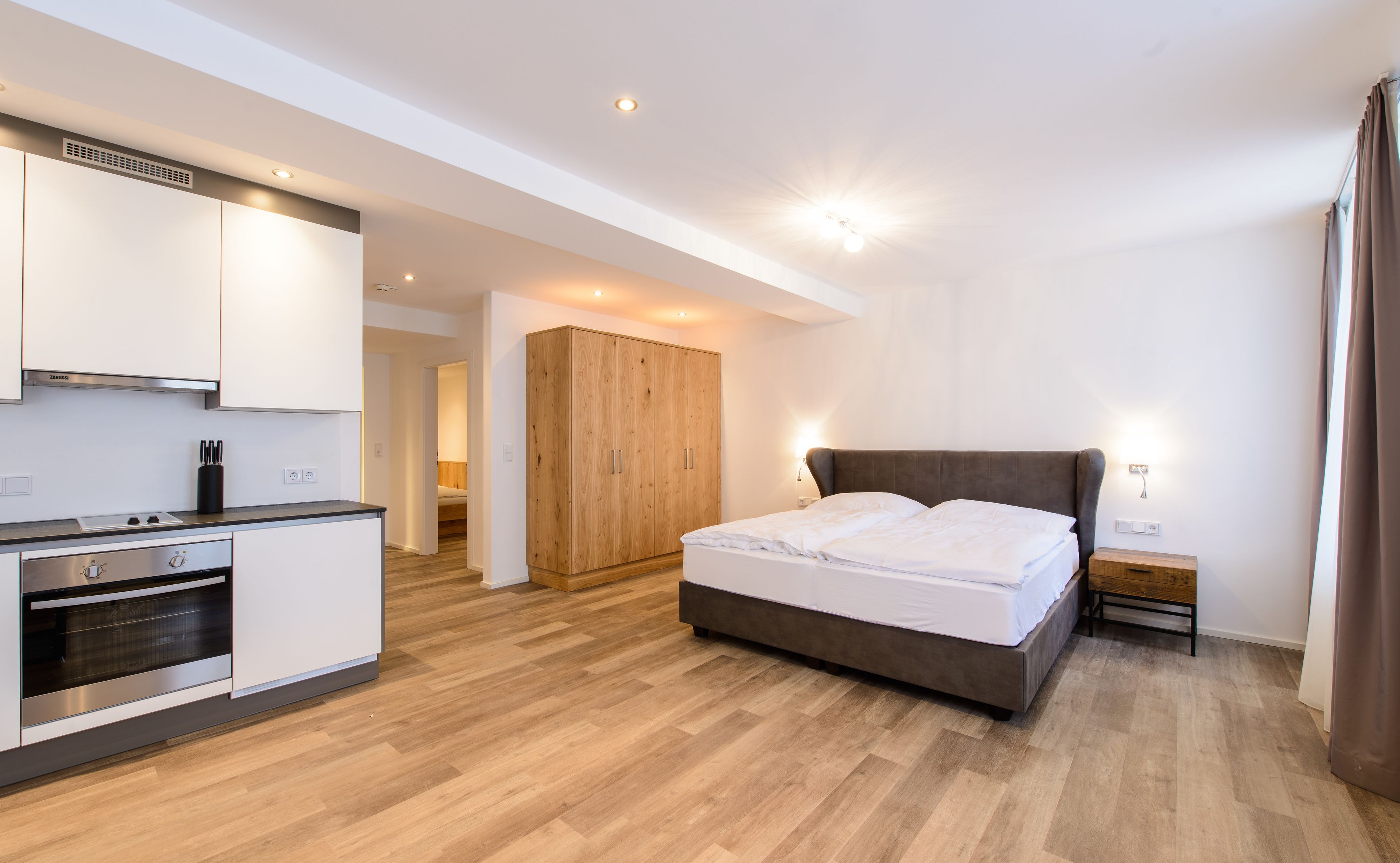 Een moderne slaapkamer met een groot bed, een houten garderobe, een kitchenette en een badkamer die door een open deur zichtbaar is.