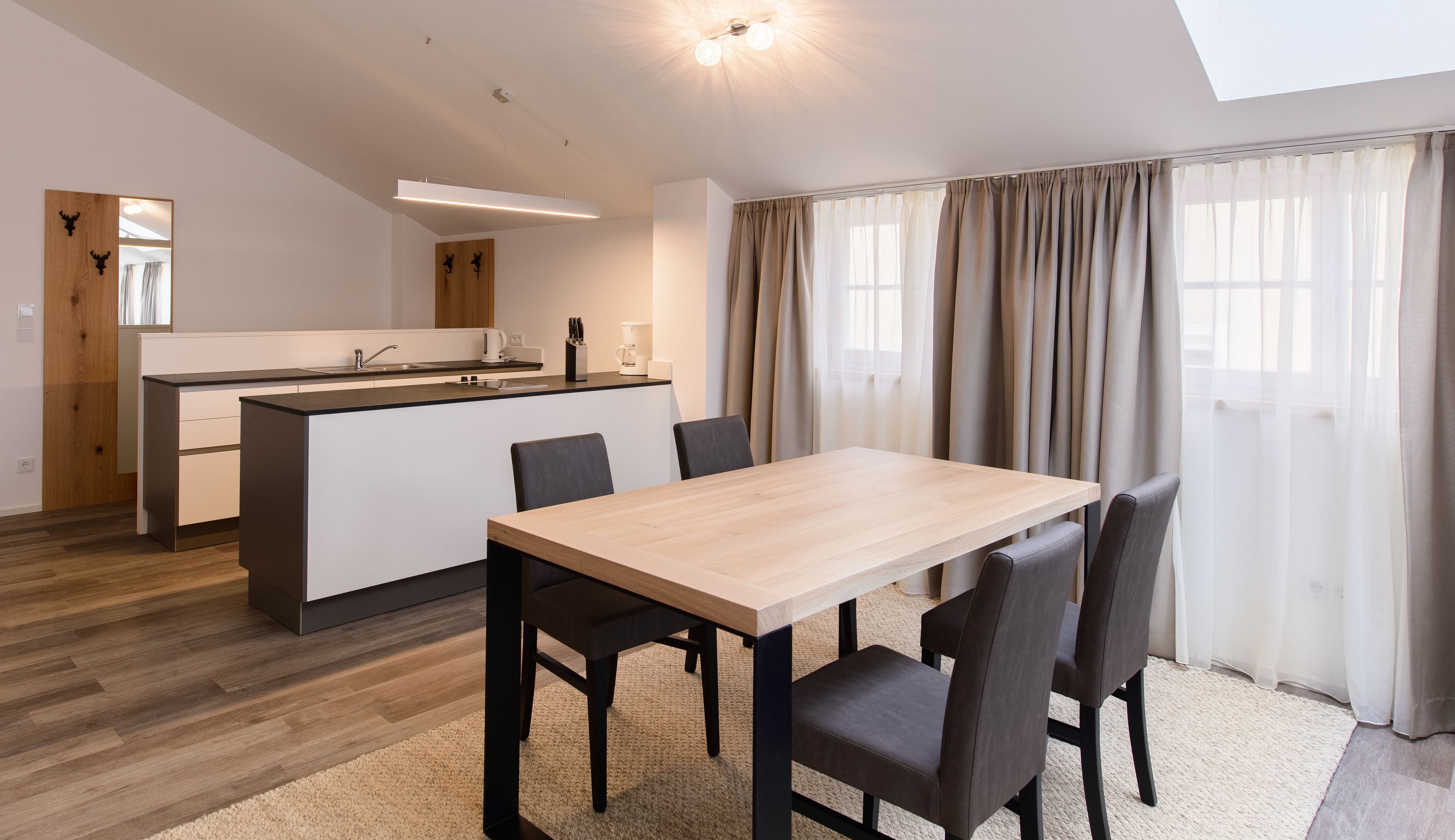 A modern kitchen with a central island, a dining area with a wooden table and chairs, and a large window with curtains.