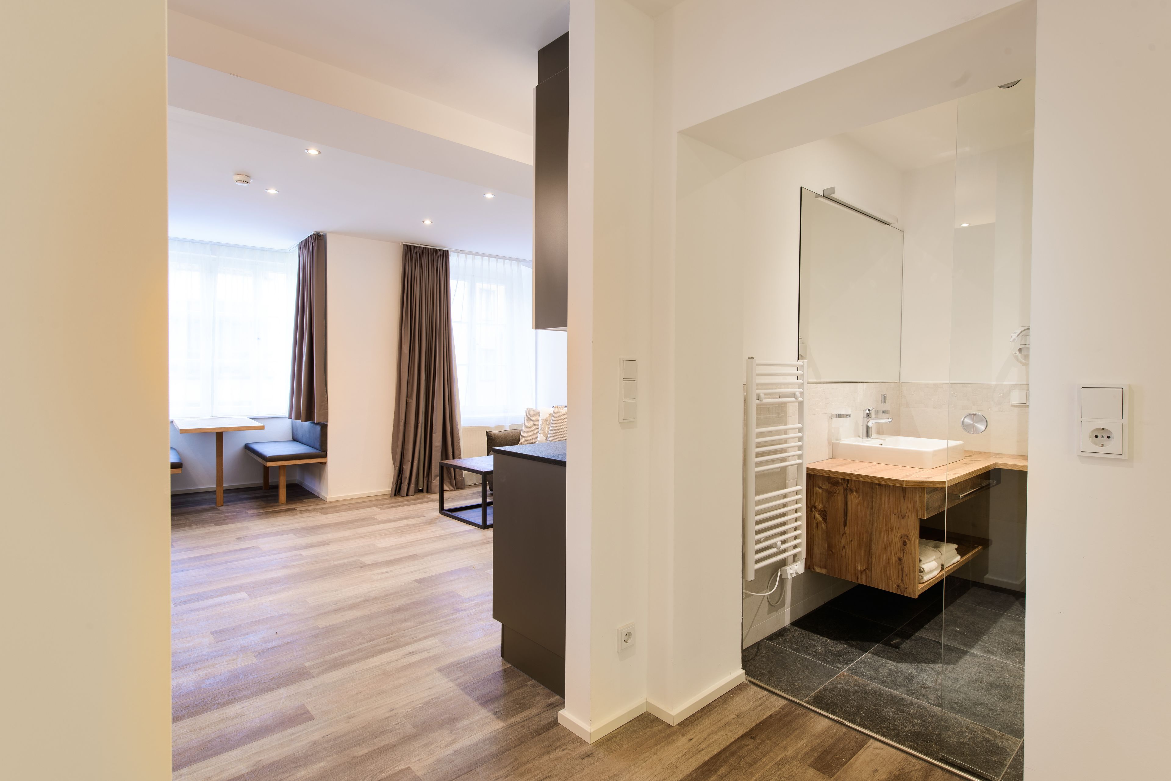 A modern hotel room with a wooden floor, a small seating area with a table and chairs, a large mirror above the sink, and a view of the outdoors through a window.