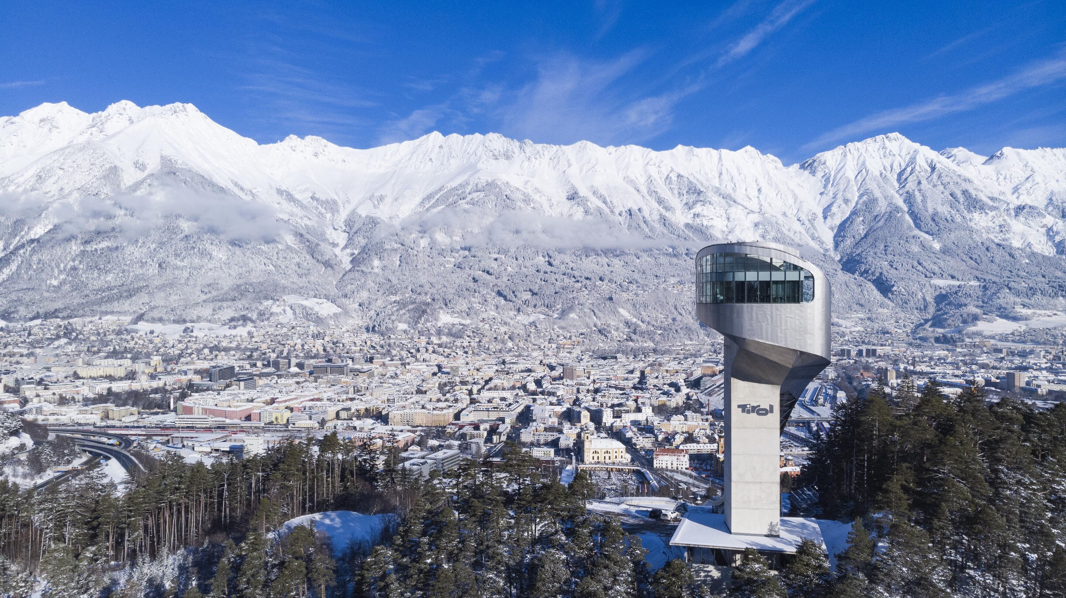 A modern tower with a glass observation deck overlooking a snow-covered city and mountains.