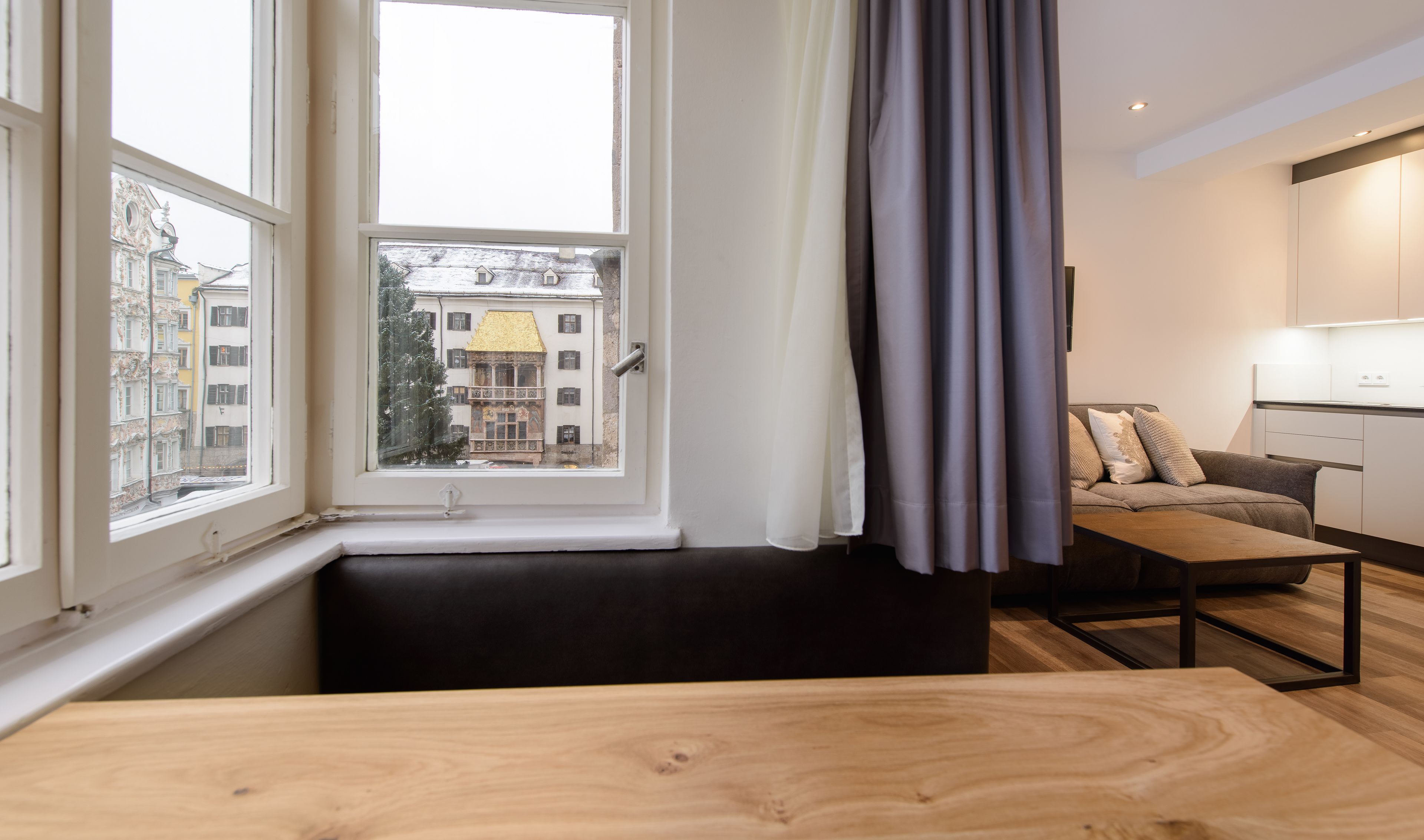 A modern living room with a large window, a sofa, a wooden table, and a kitchen area.