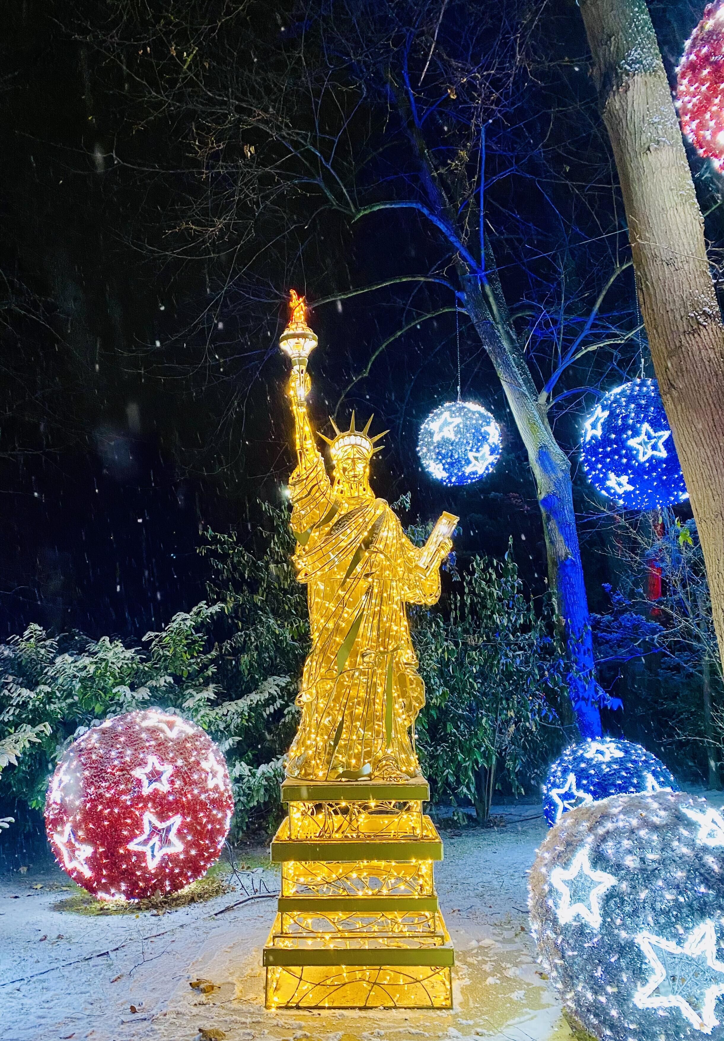 Une statue dorée représentant une figure tenant une torche, entourée de sphères illuminées et de décorations recouvertes de neige.
