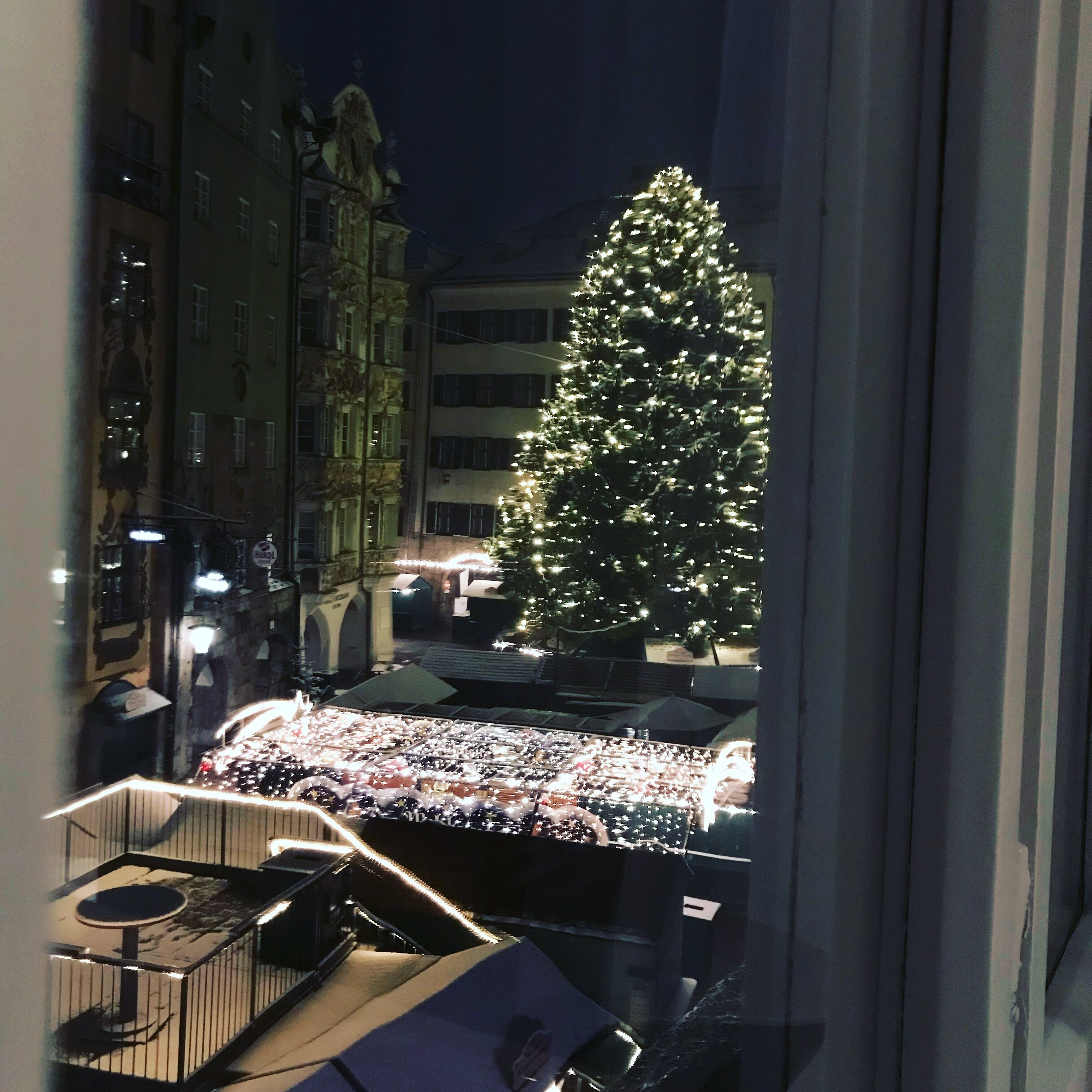 A view of a Christmas tree decorated with lights, a balcony with a table and chairs, and a building with classical architecture in the background.