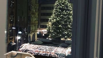 A view of a Christmas tree decorated with lights, a balcony with a table and chairs, and a building with classical architecture in the background.