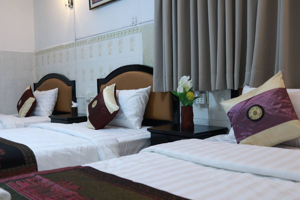 A hotel room with three single beds, each with a headboard, bedside tables, and pillows. There is a nightstand with a vase of flowers between the beds.