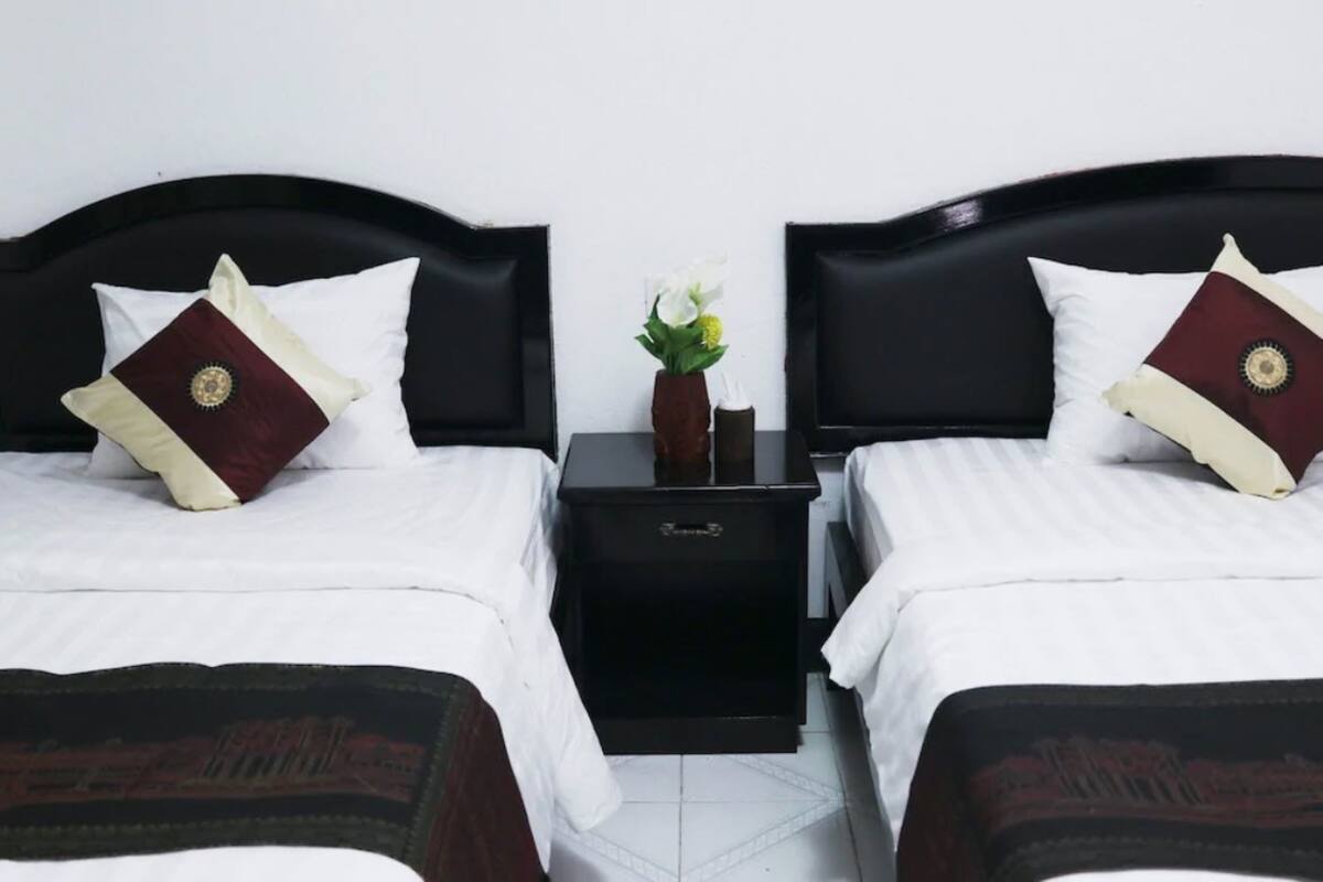 A hotel room with two beds, each with a black headboard, white bedding, and decorative pillows. A small black nightstand with a vase of flowers is between the beds.