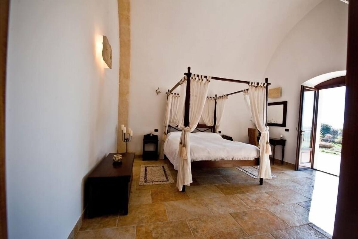 Un lit à baldaquin à quatre poteaux, agrémenté de draps blancs, une petite table de chevet avec des bougies et une vue sur l’extérieur grâce à une porte vitrée.