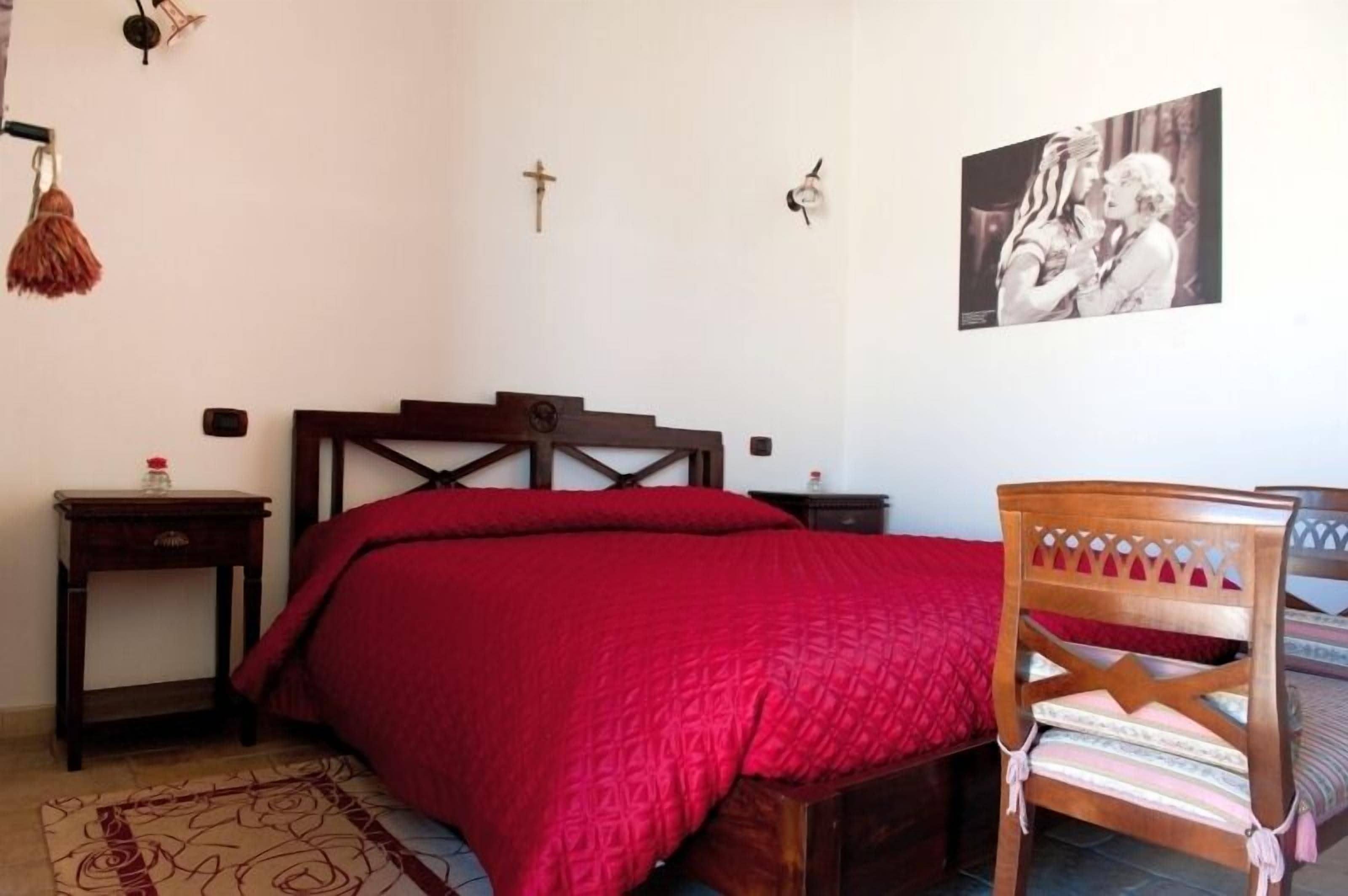Un dormitorio con una cama de madera, un edredón rojo, una silla de madera, una mesita con una botella y una cruz colgada en la pared.