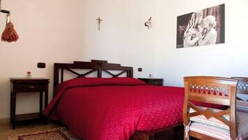 Un dormitorio con una cama de madera, un edredón rojo, una silla de madera, una mesita con una botella y una cruz colgada en la pared.